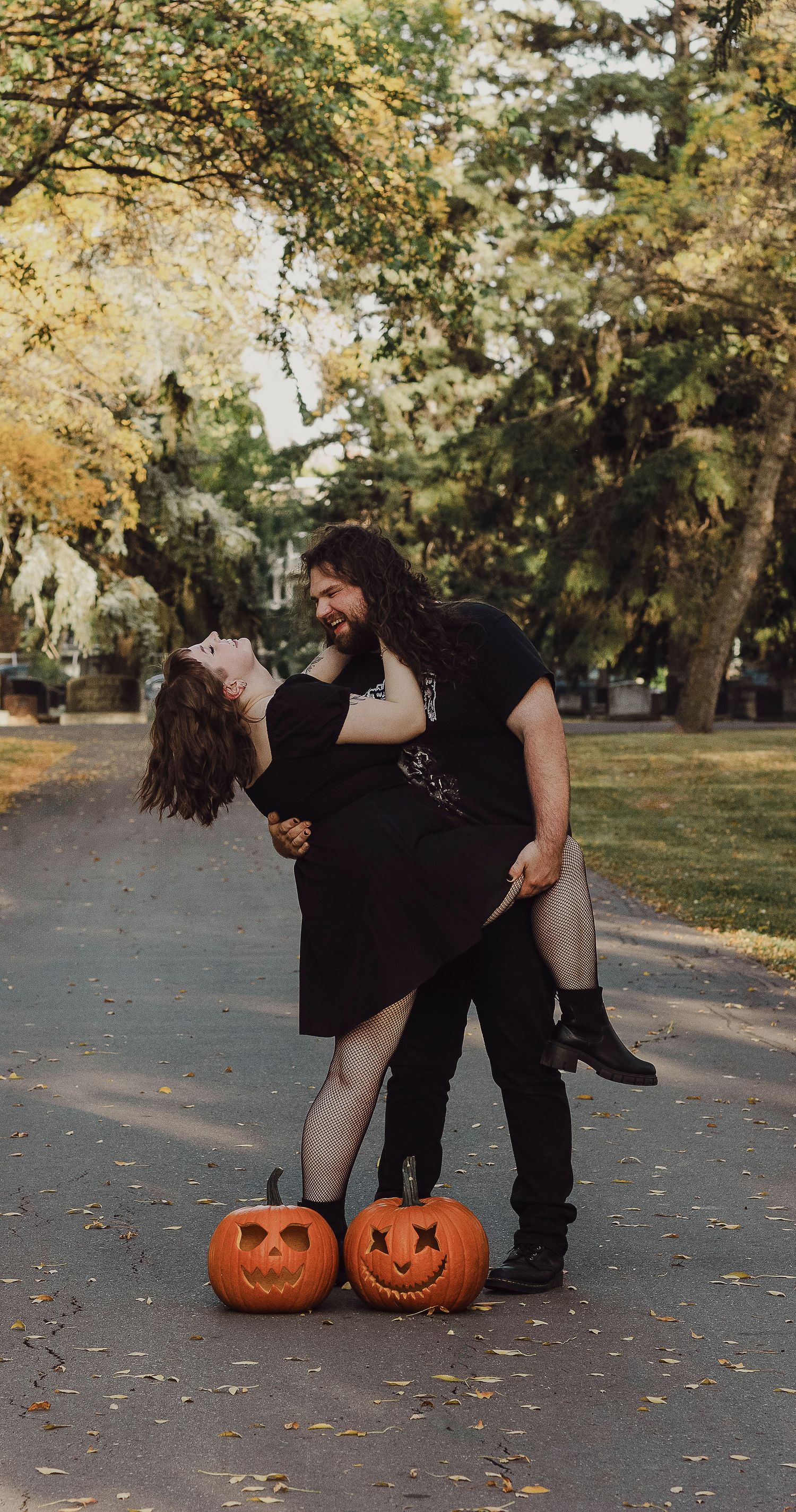 Two carved jack-o'-lanterns sit on a leaf-covered path while a couple in black clothing shares a romantic moment in autumn.