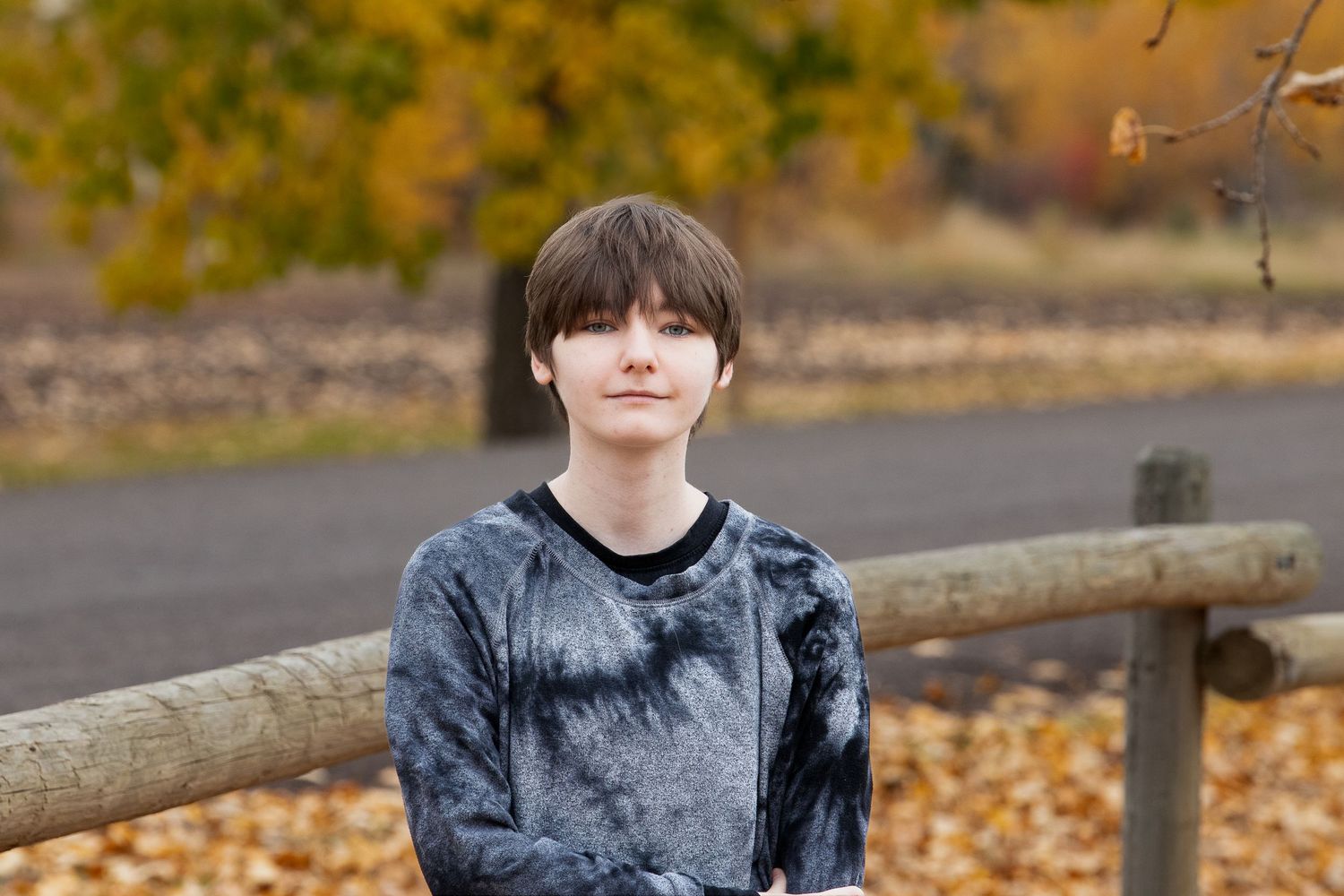 Person wearing a dark gray sweater sits by a wooden fence during autumn with yellow leaves in the background.