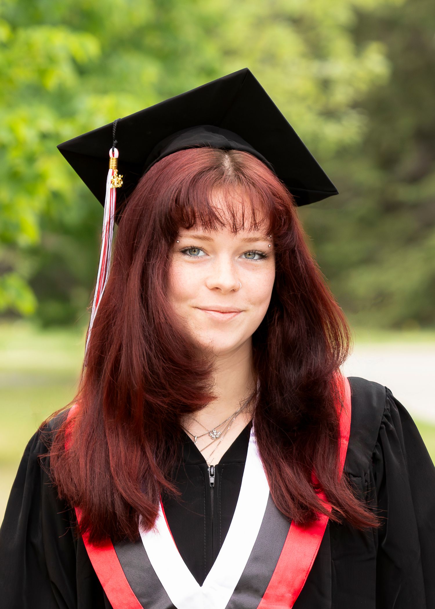 Graduate in black cap and gown with red trim against green foliage background.