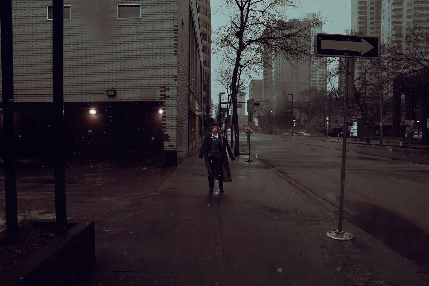 A person walks down a dimly lit urban sidewalk at dusk with high-rise buildings in the background.