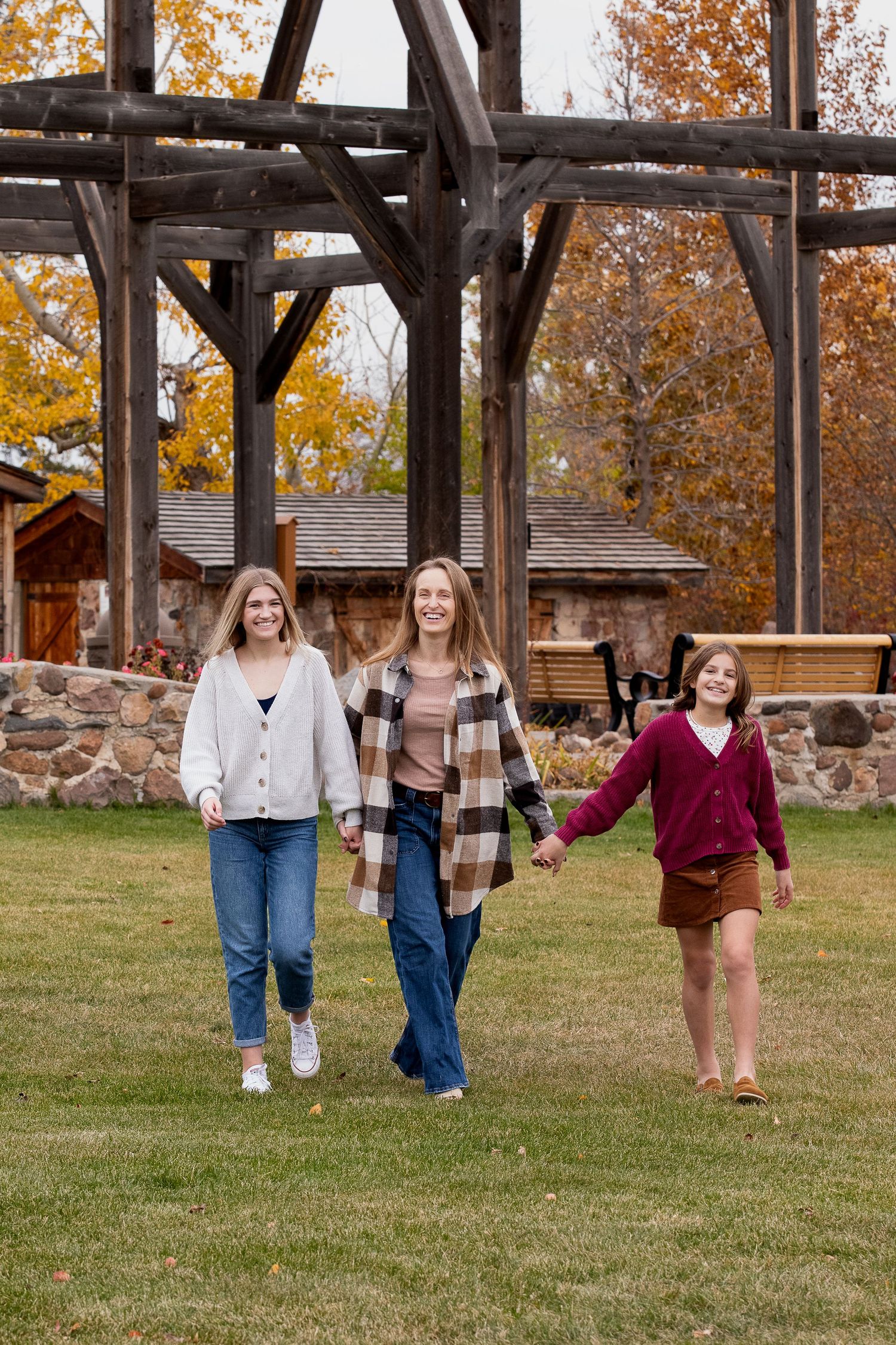 Three people walk together on a grassy lawn beneath a rustic wooden pergola with autumn trees in the background.