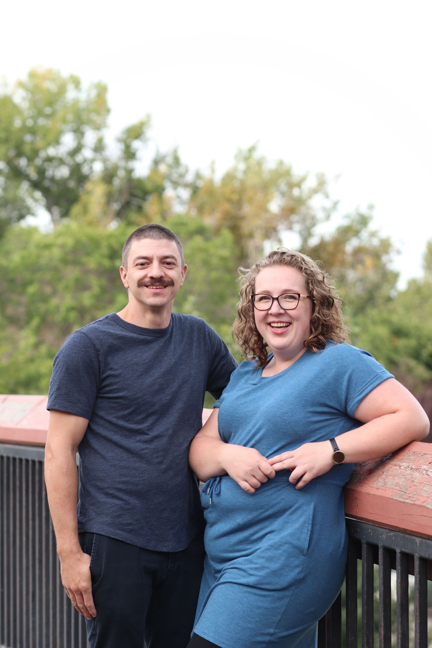 A couple smiles together while standing on an outdoor deck with trees visible in the background.