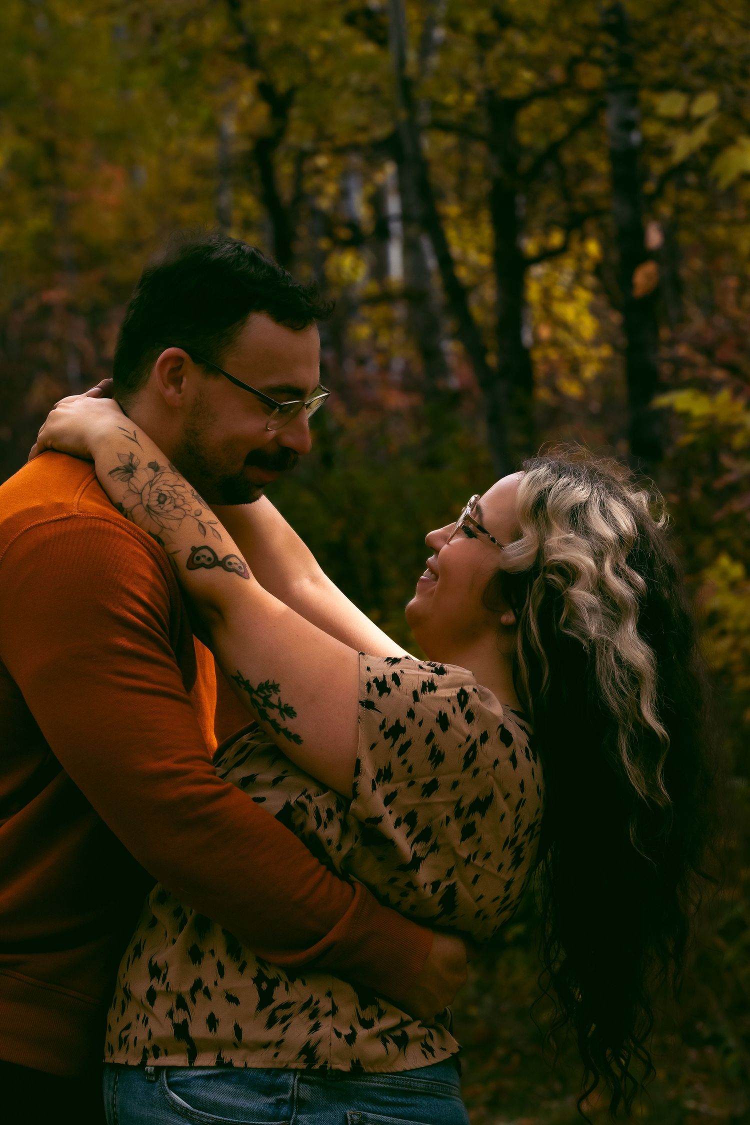 A romantic autumn photoshoot shows a couple embracing and laughing together in a forest with golden fall foliage.