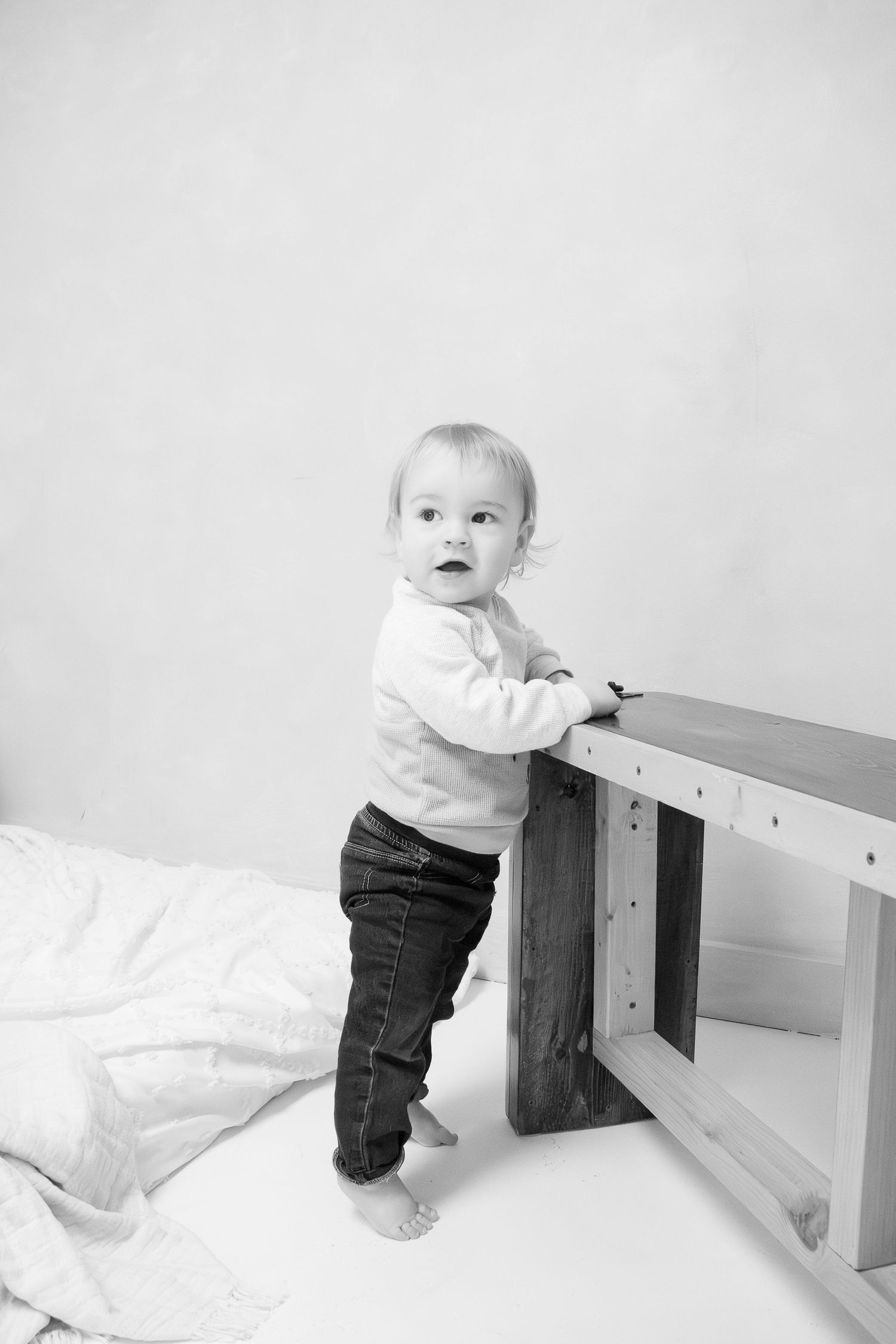 Black and white photo of a toddler in a white top and jeans standing at a wooden bench structure.
