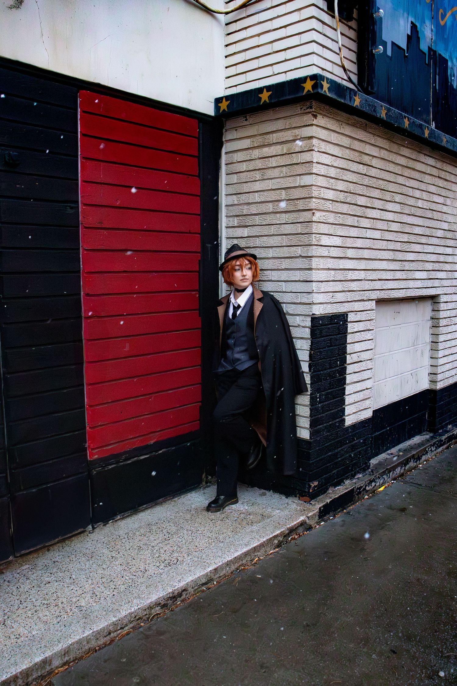 Someone in vintage attire stands against a wall with contrasting red and white panels on a rainy city street.