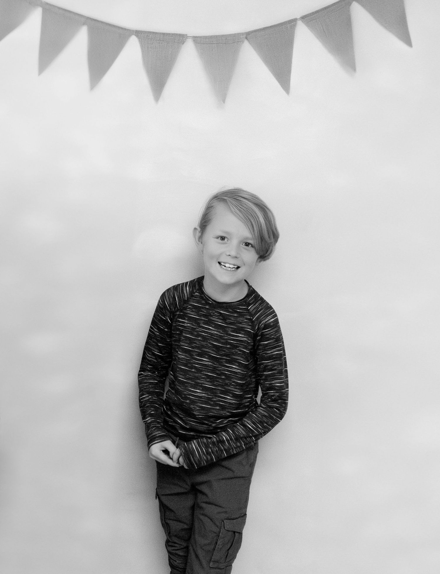 Black and white portrait of a child in a striped sweater standing against a wall with decorative bunting banner.
