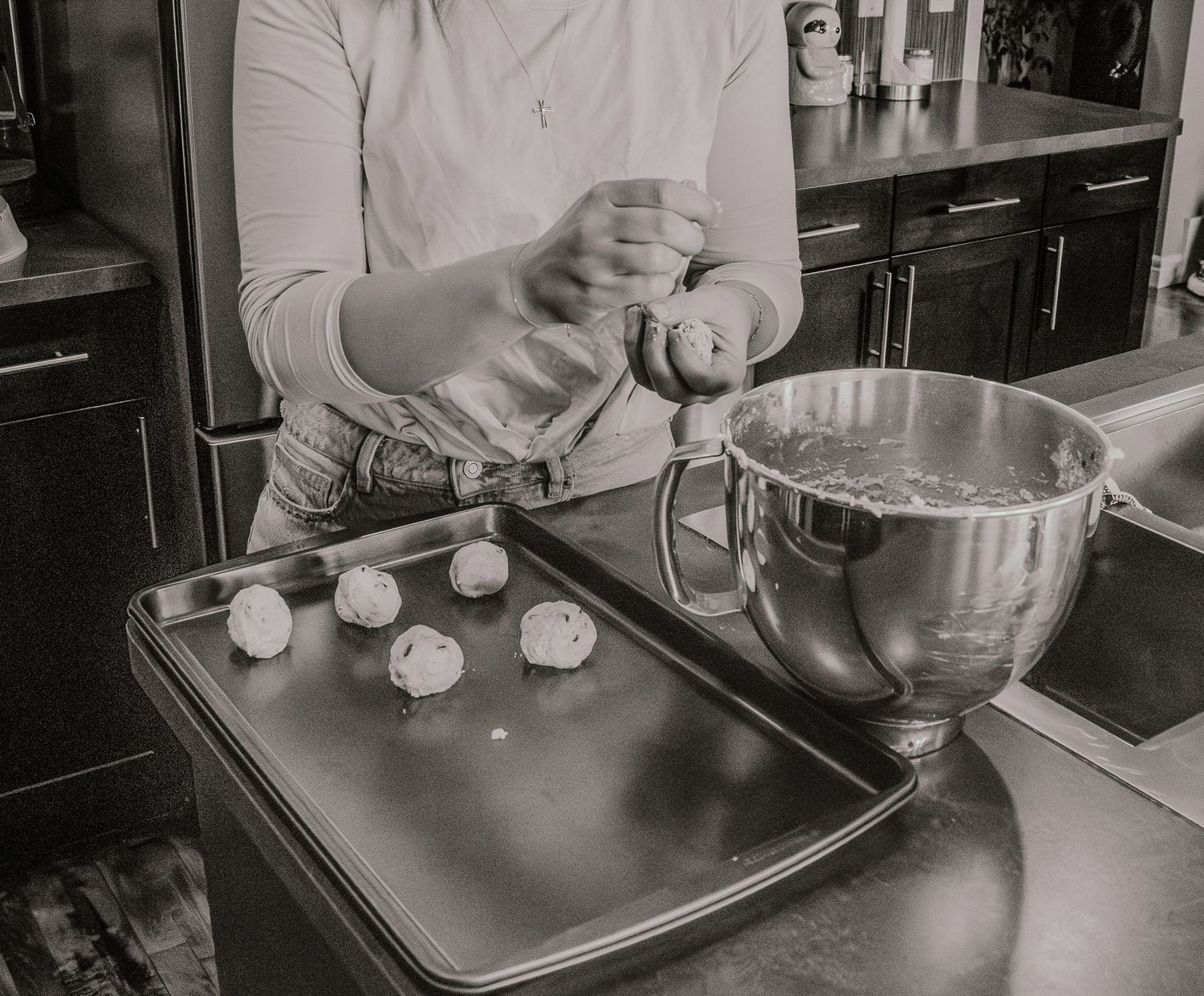 A baker preparing small dough portions on a baking sheet next to a large mixing bowl in a black and white photograph.