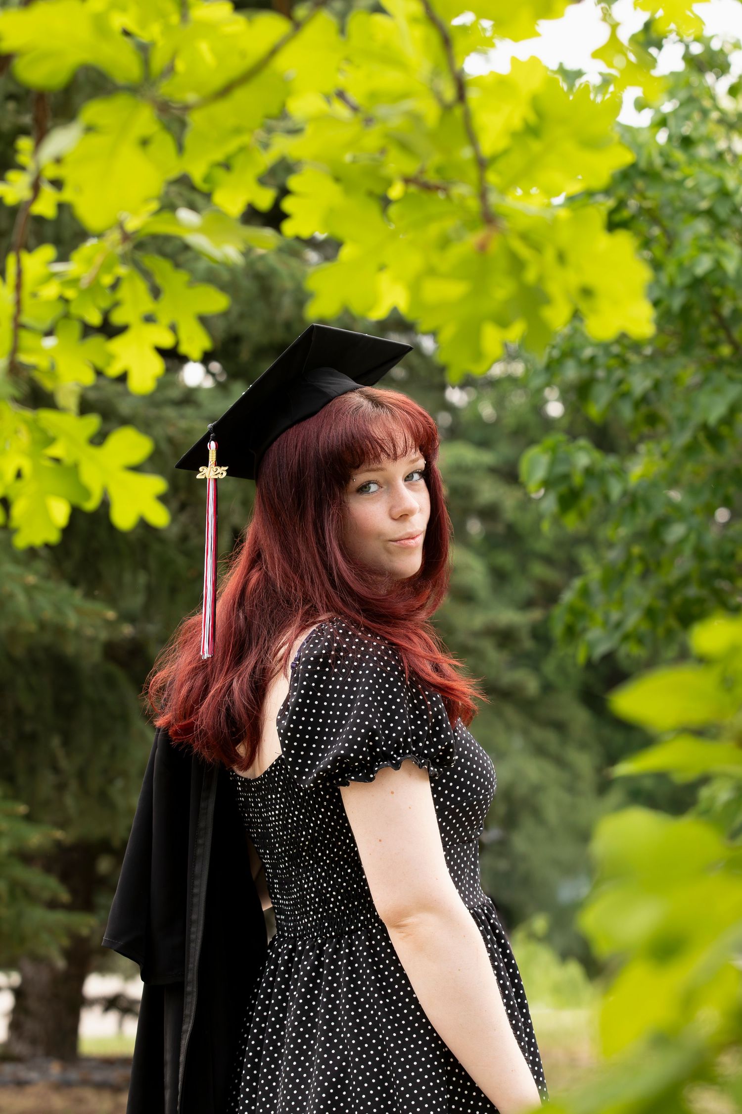 Graduate in black cap and gown with polka dot dress poses in front of green leaves.