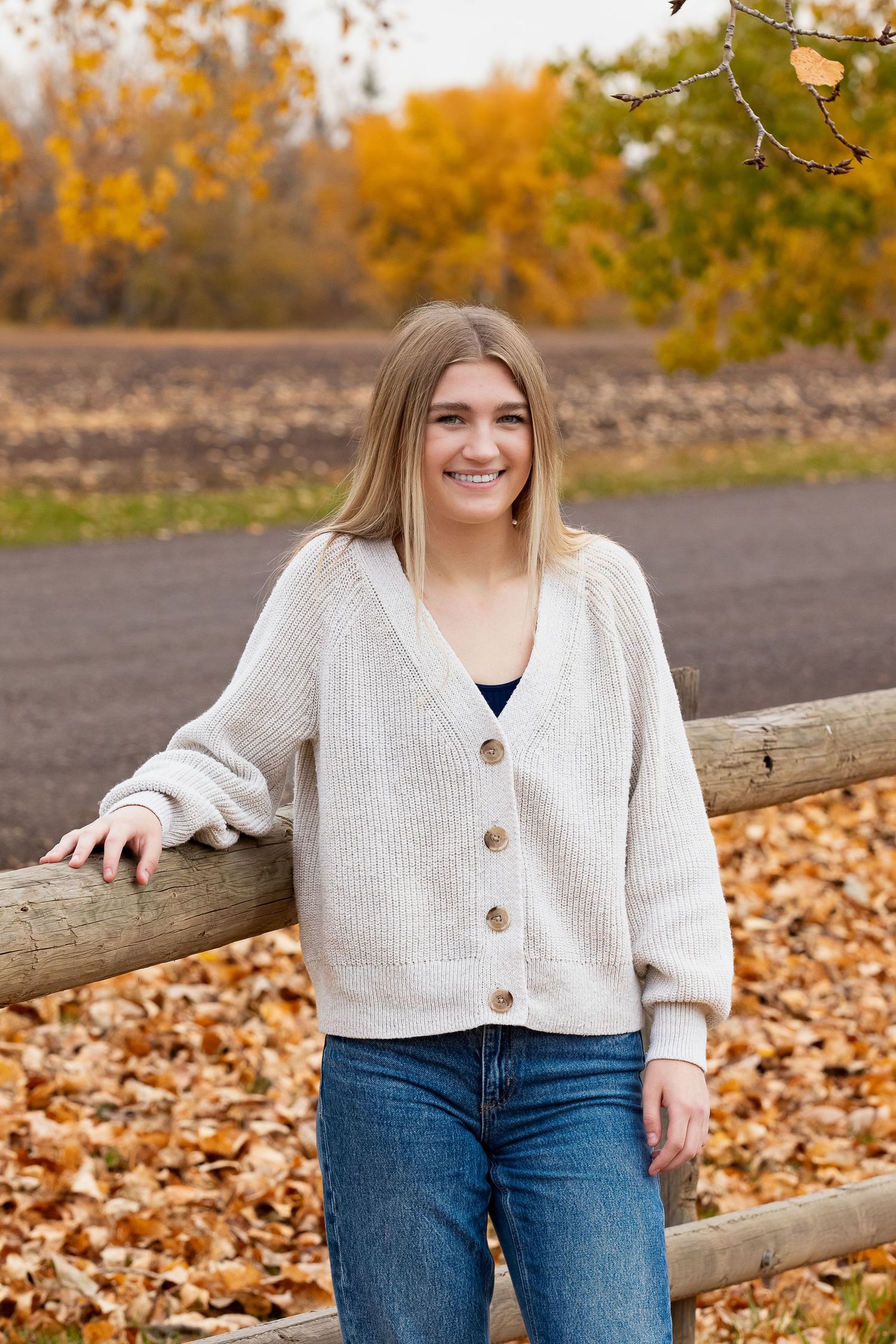 Someone in a cozy cream cardigan and jeans stands by a rustic wooden fence surrounded by autumn foliage.