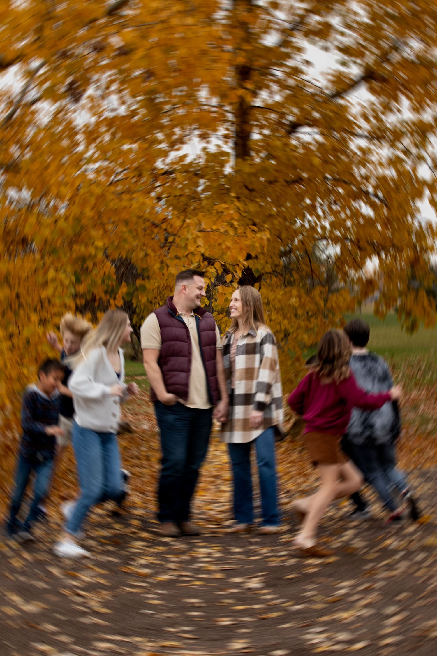 A group stands together under vibrant fall foliage while autumn leaves swirl around them in a creative motion blur effect.