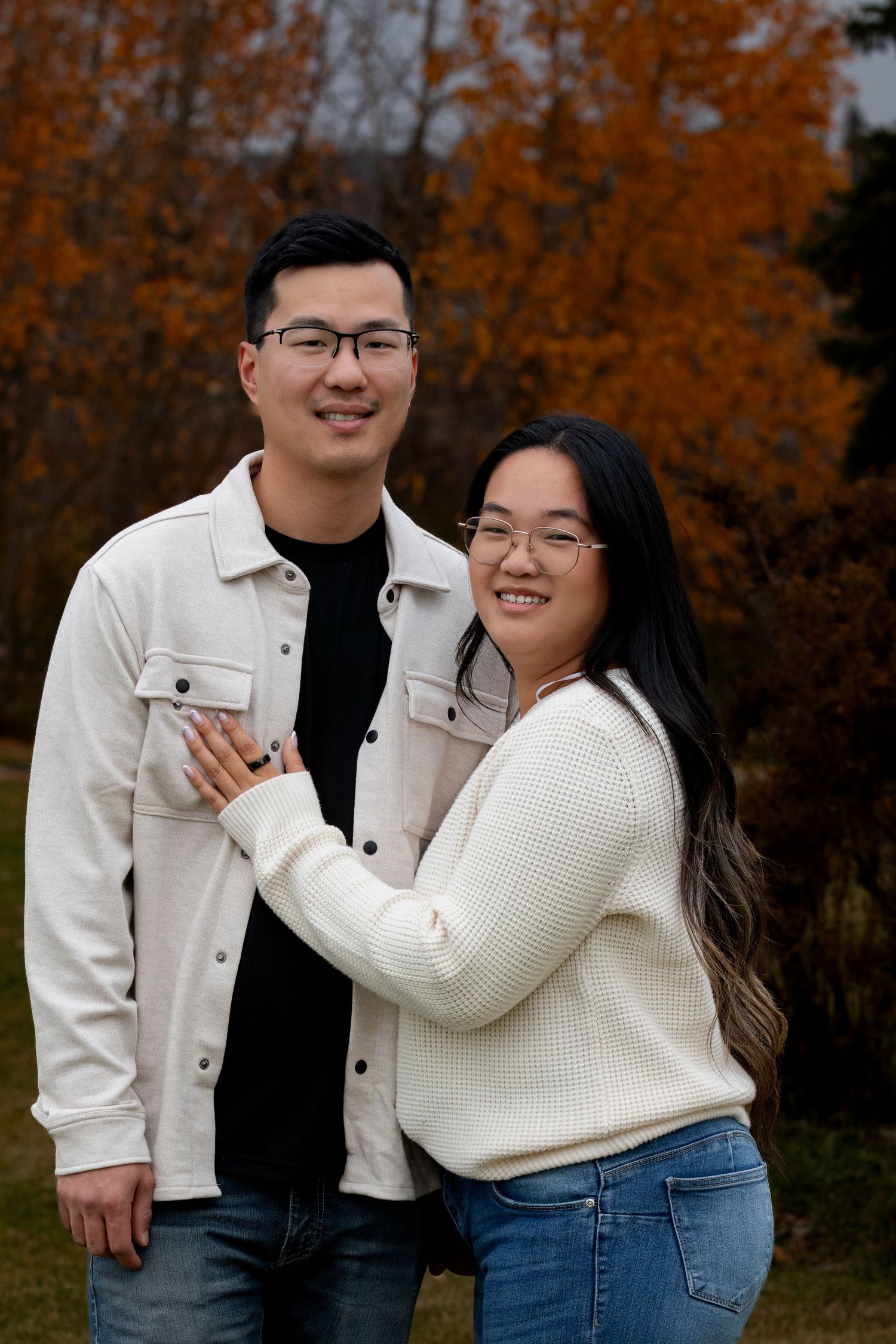 A loving couple poses together outdoors against fall foliage wearing cozy sweaters and smiling warmly.