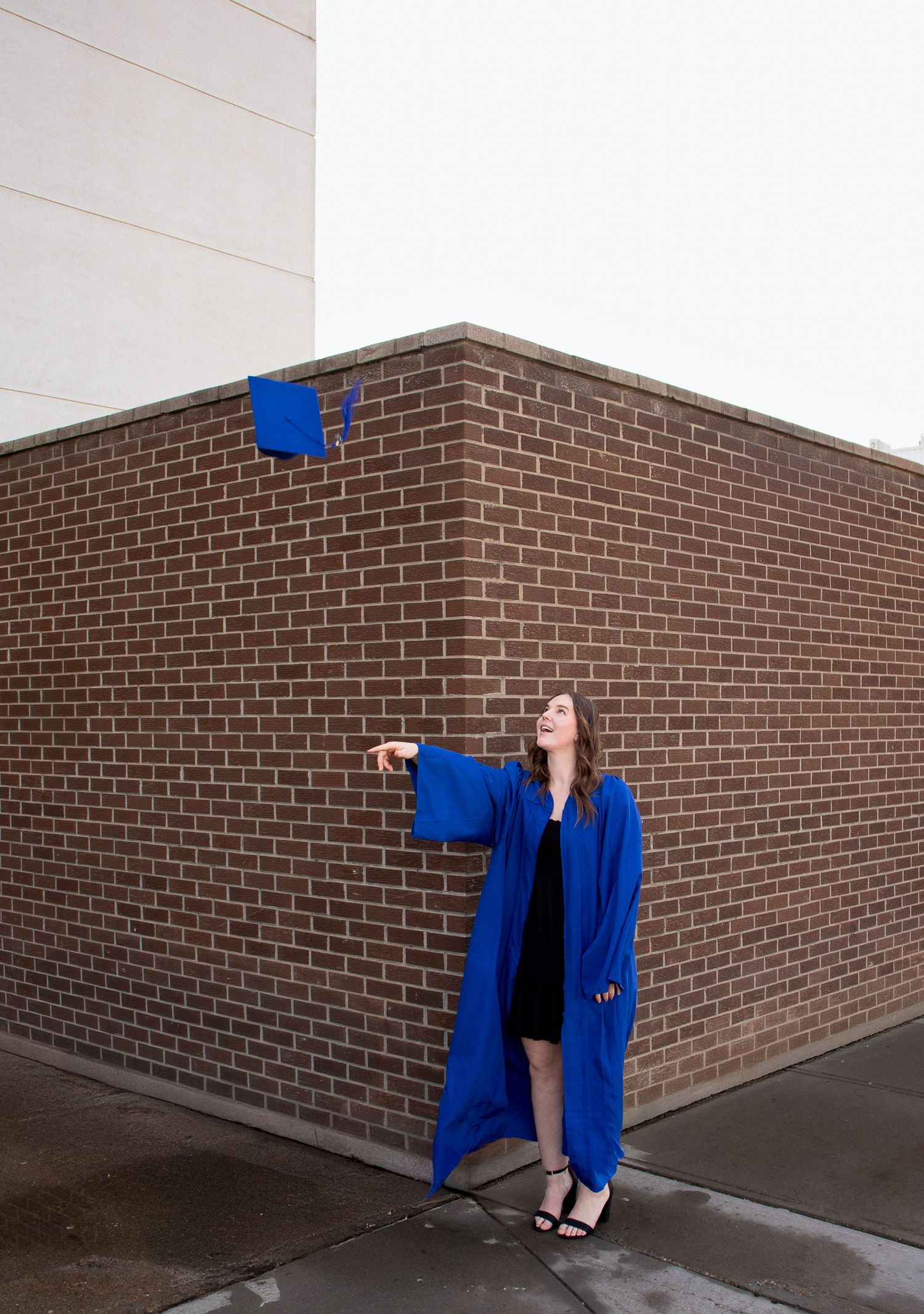 Graduate in blue cap and gown poses playfully against brick wall in graduation photo series.