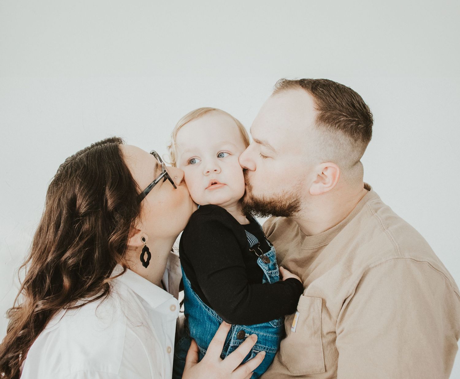 Two parents share a tender moment kissing their baby dressed in black against a light gray studio backdrop.