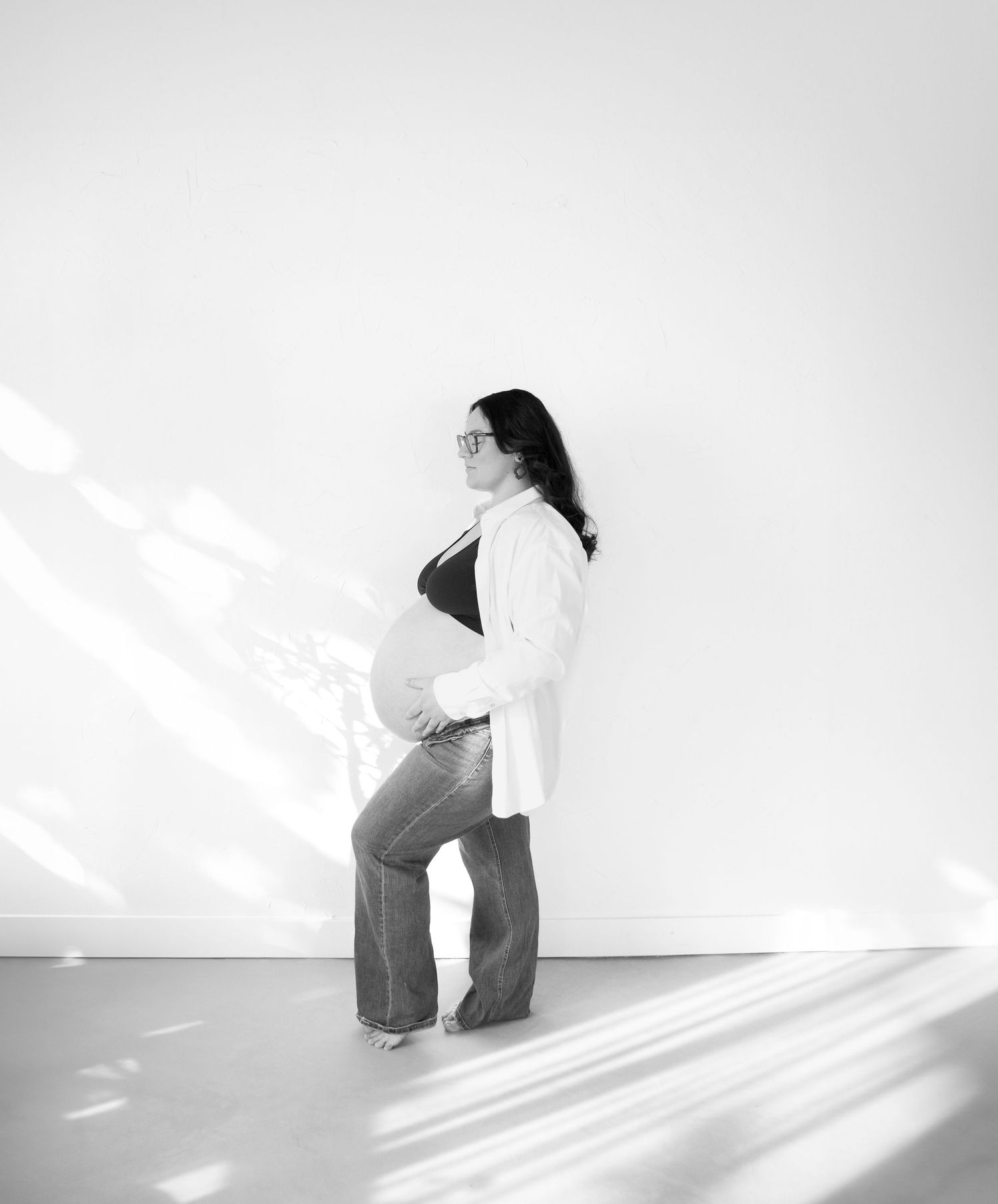 Black and white portrait of pregnant woman posing against a white wall with dramatic diagonal shadows streaming across the scene.
