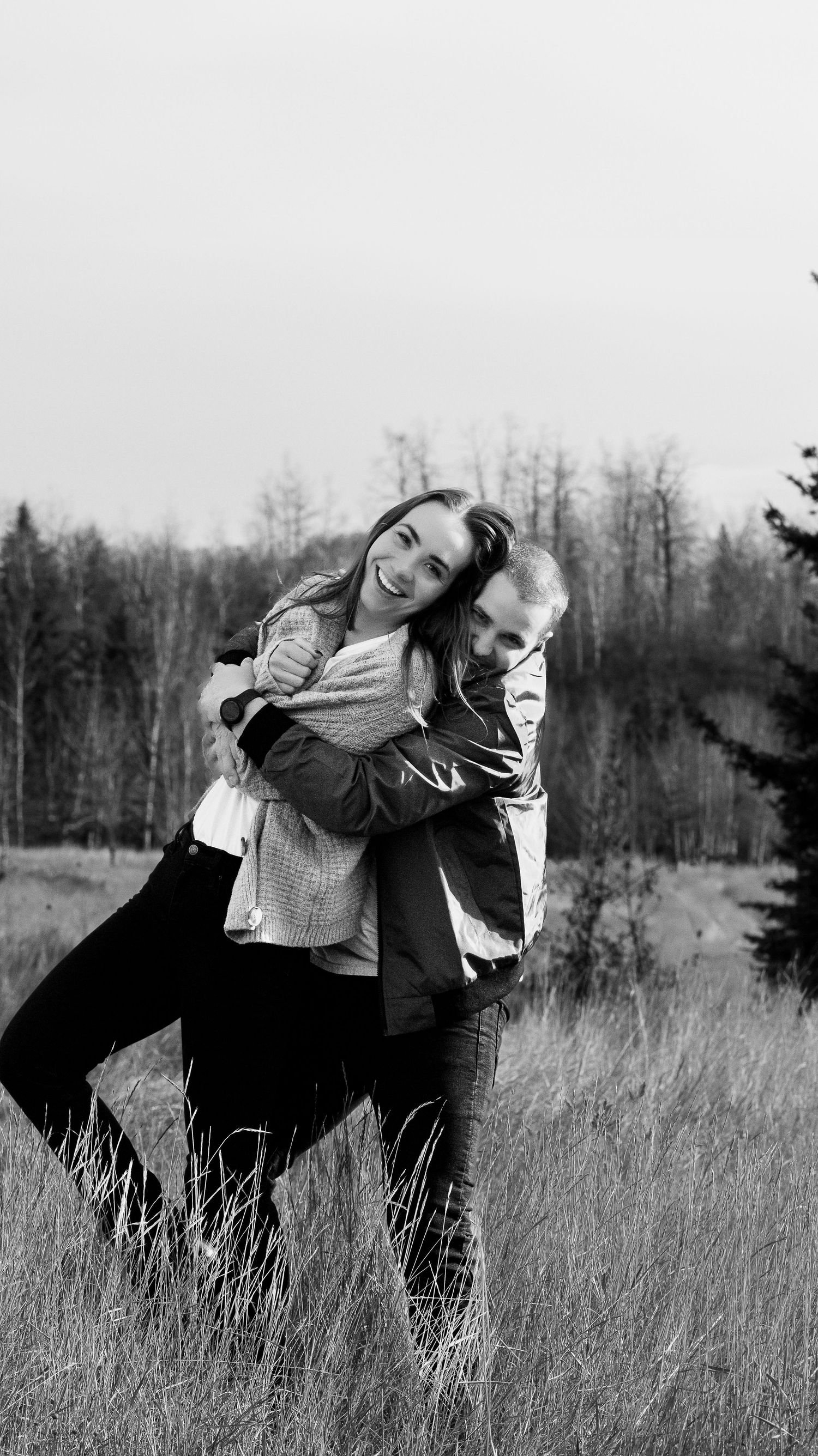 A couple embraces playfully in a black and white photo taken in a field with trees in the background.