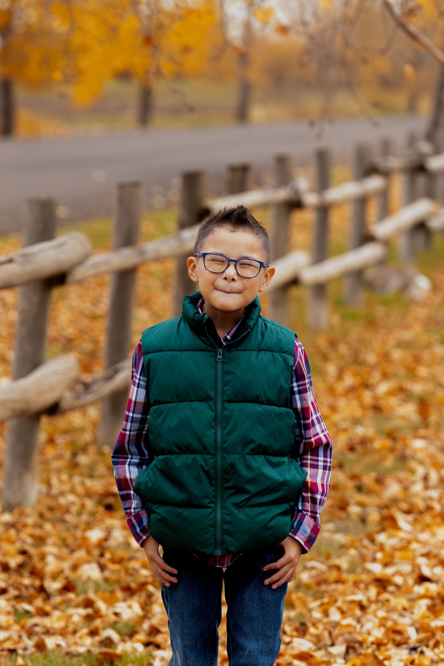 A child in a green vest and plaid shirt stands smiling outdoors among fallen autumn leaves and wooden fencing.