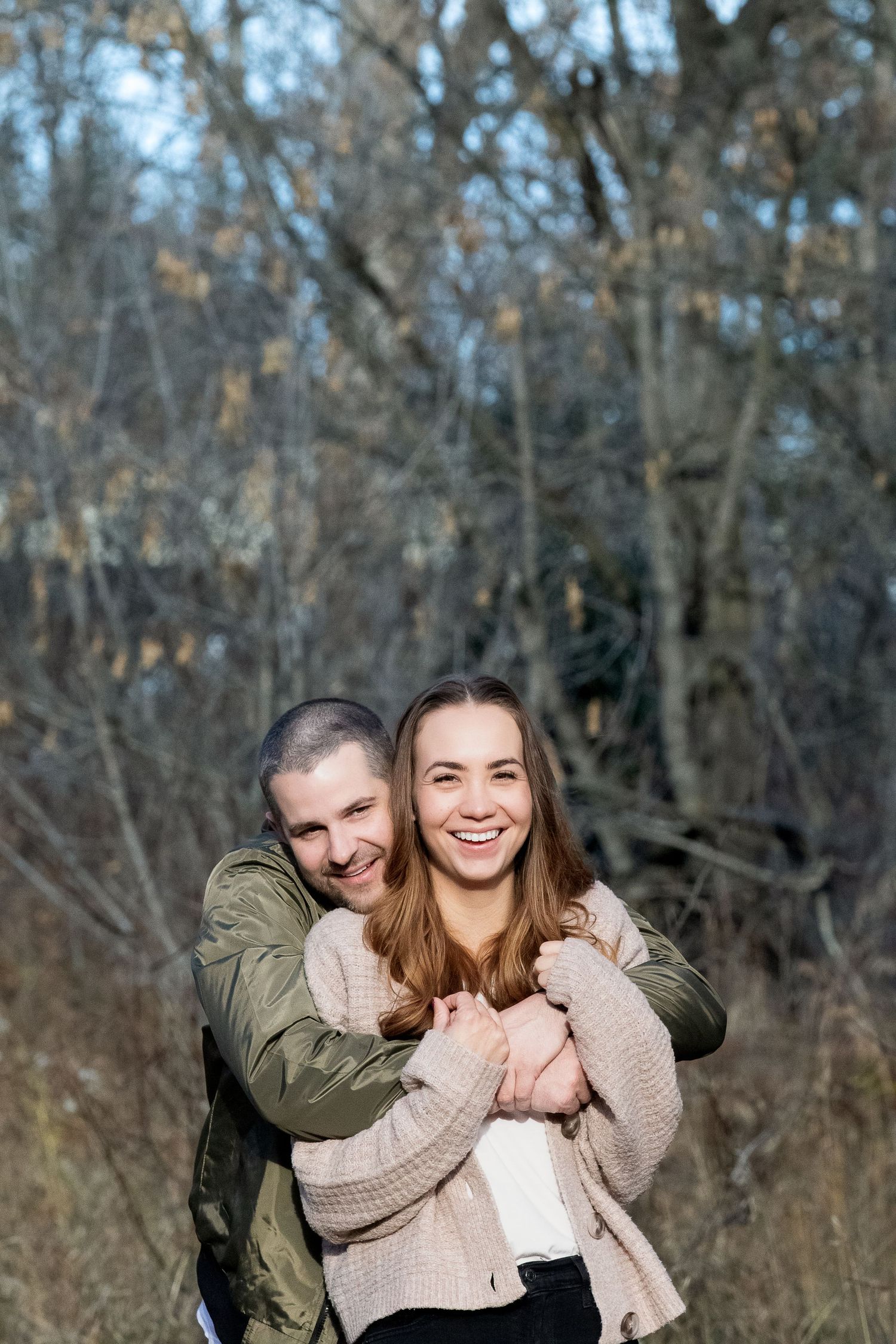 A couple embraces and laughs together in casual sweaters while standing in front of bare autumn trees in nature.