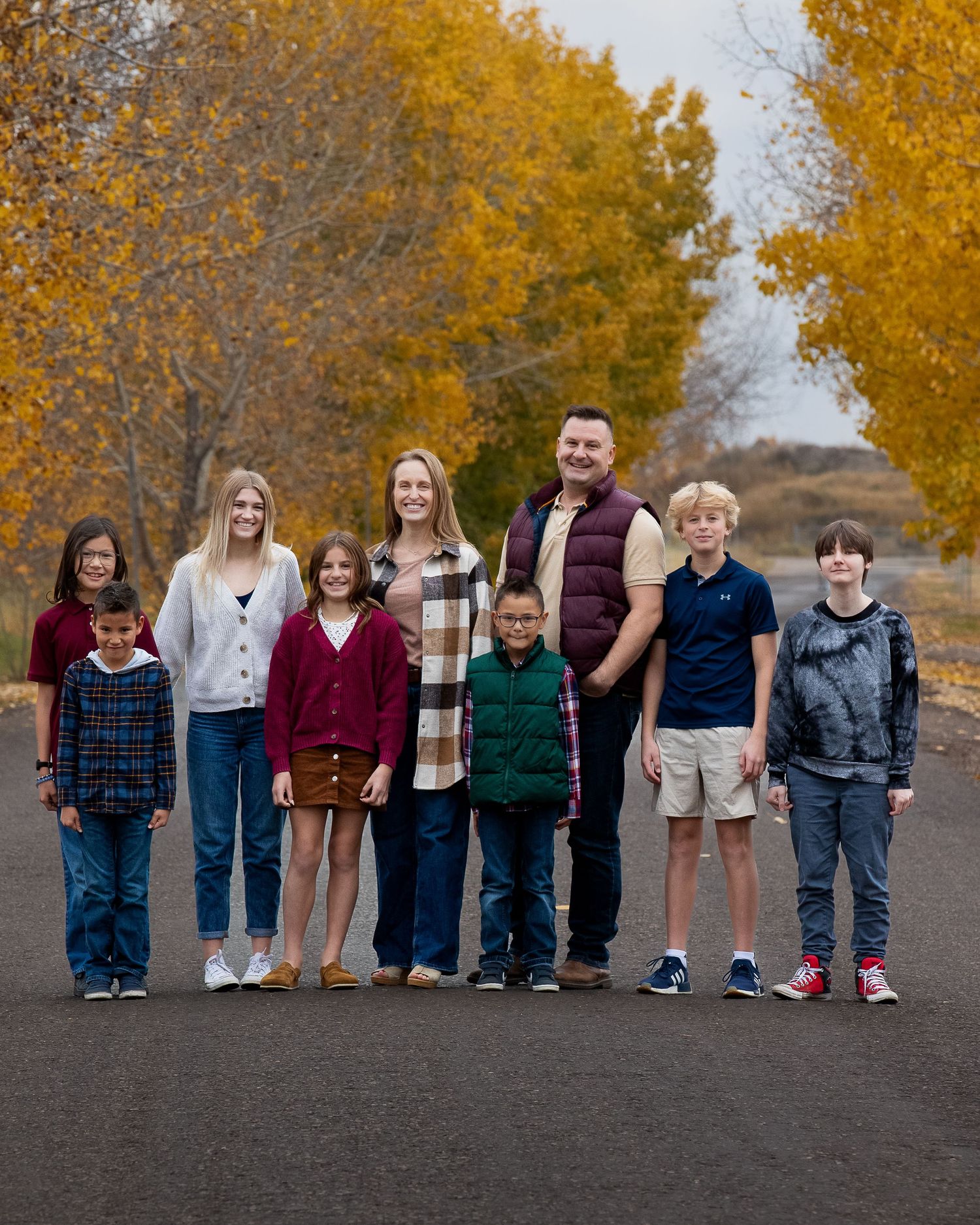 A mixed family together on an autumn road lined with golden trees wearing casual clothing for a family portrait.