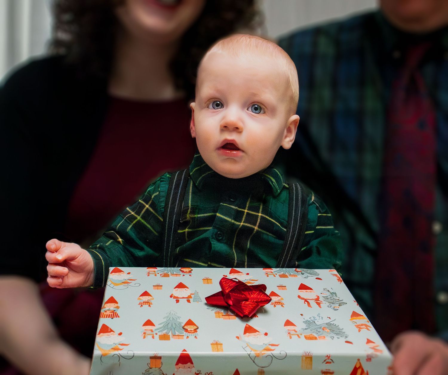 A baby in a green plaid outfit sits in front of a white Christmas gift box with a red bow on top.