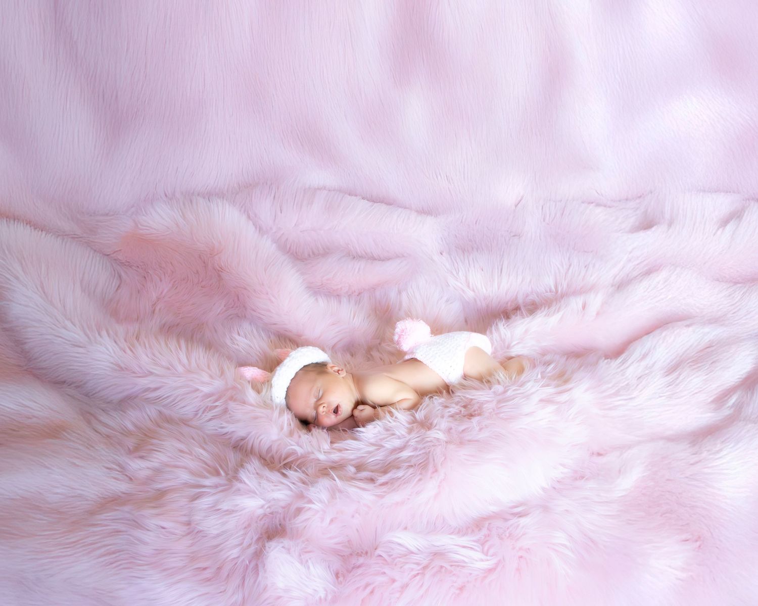 Newborn baby resting on soft pink furry blanket in dreamy light.
