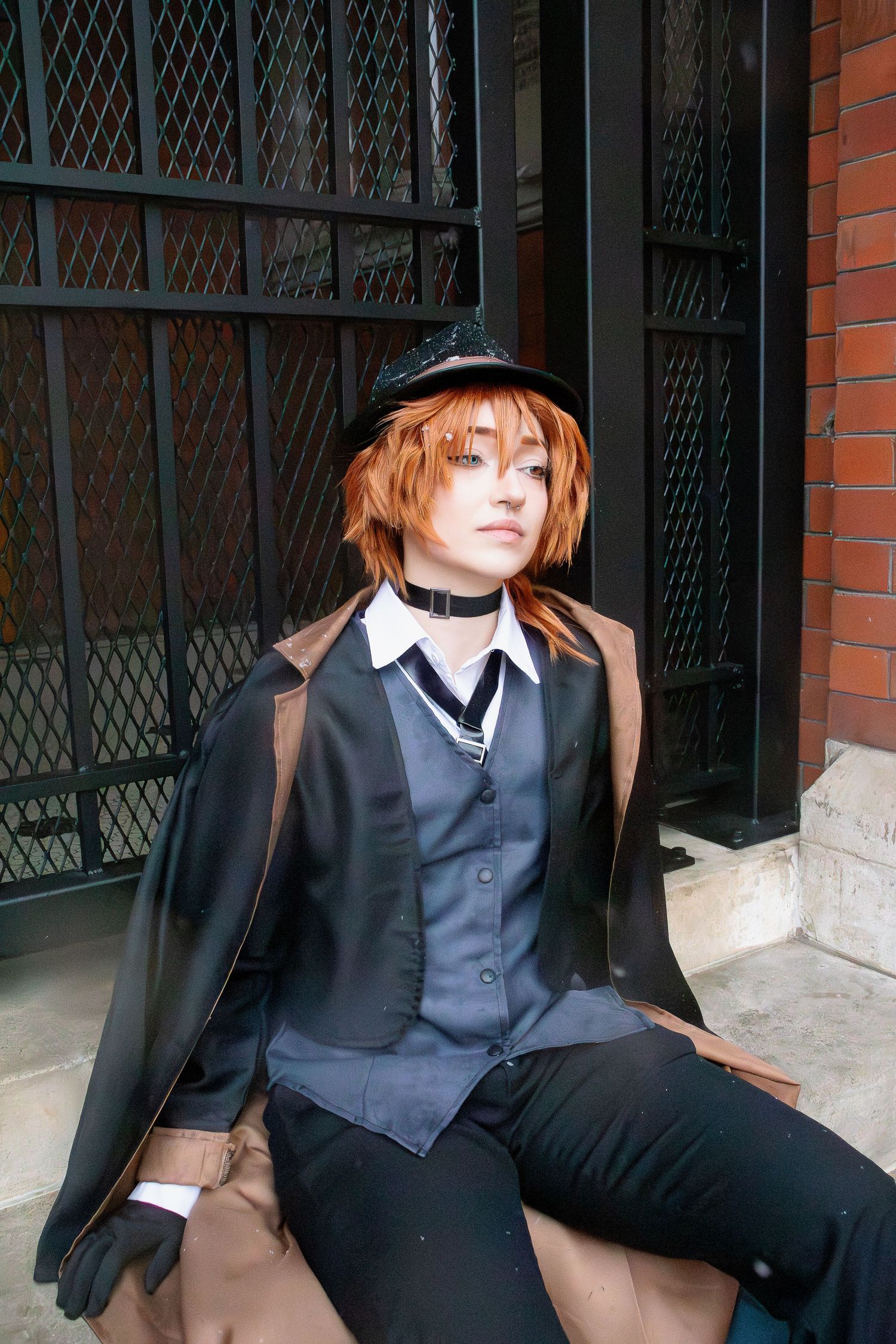 Cosplay model wearing black formal attire with vest, cape, and hat poses on stone ledge against metal grating and brick wall.