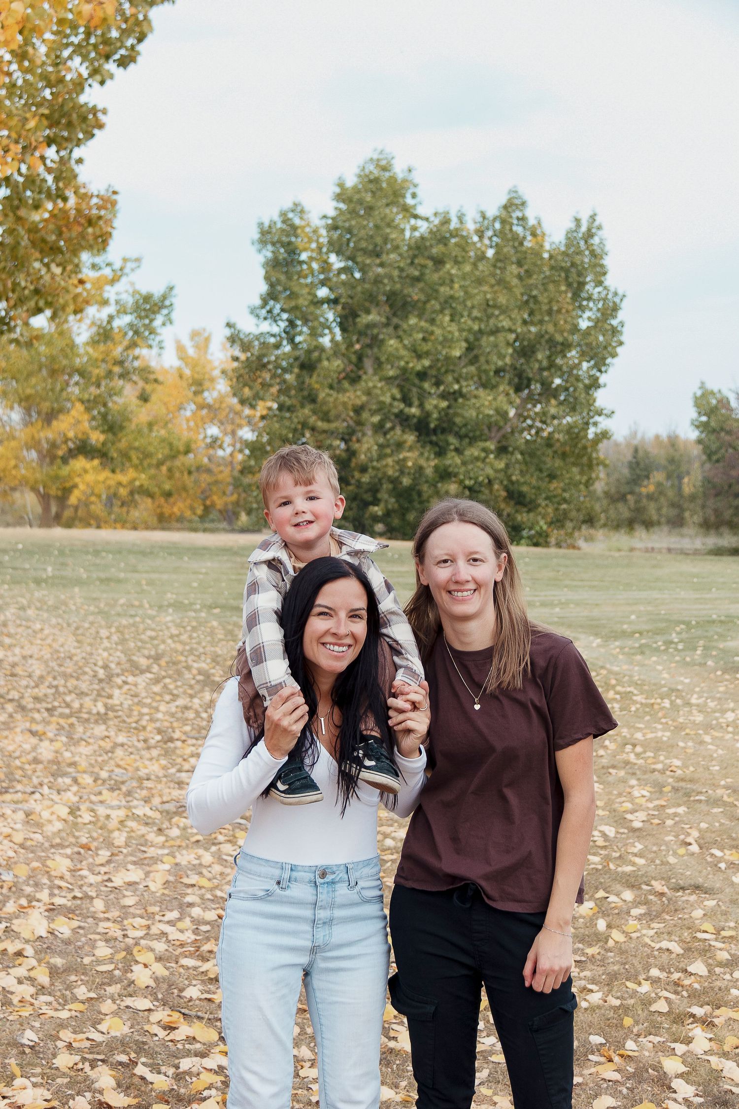 Two people laugh together outdoors on an autumn day with fallen leaves and trees in the background.