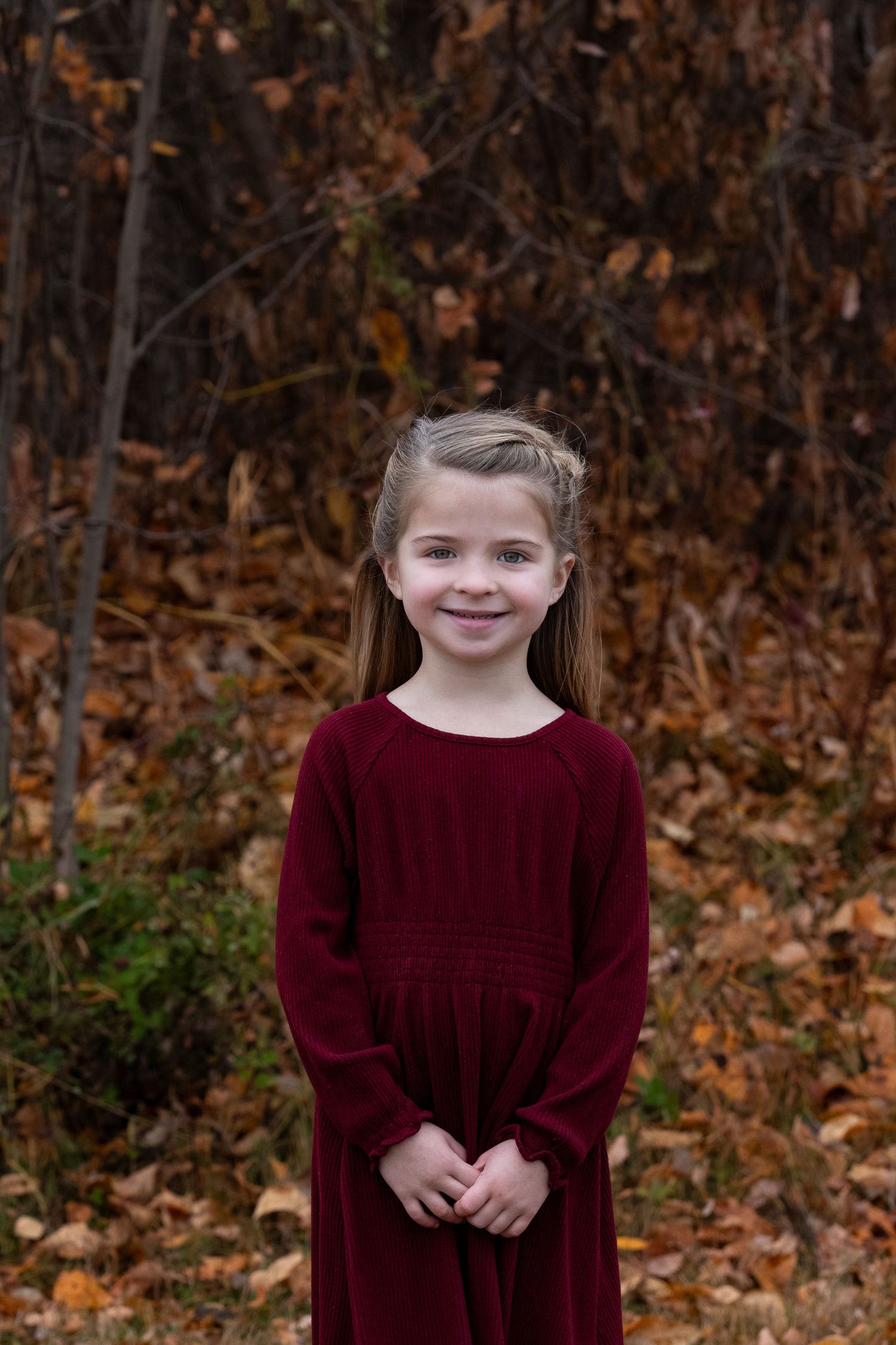 A young child in a burgundy knit dress stands in front of autumn foliage and fallen leaves.