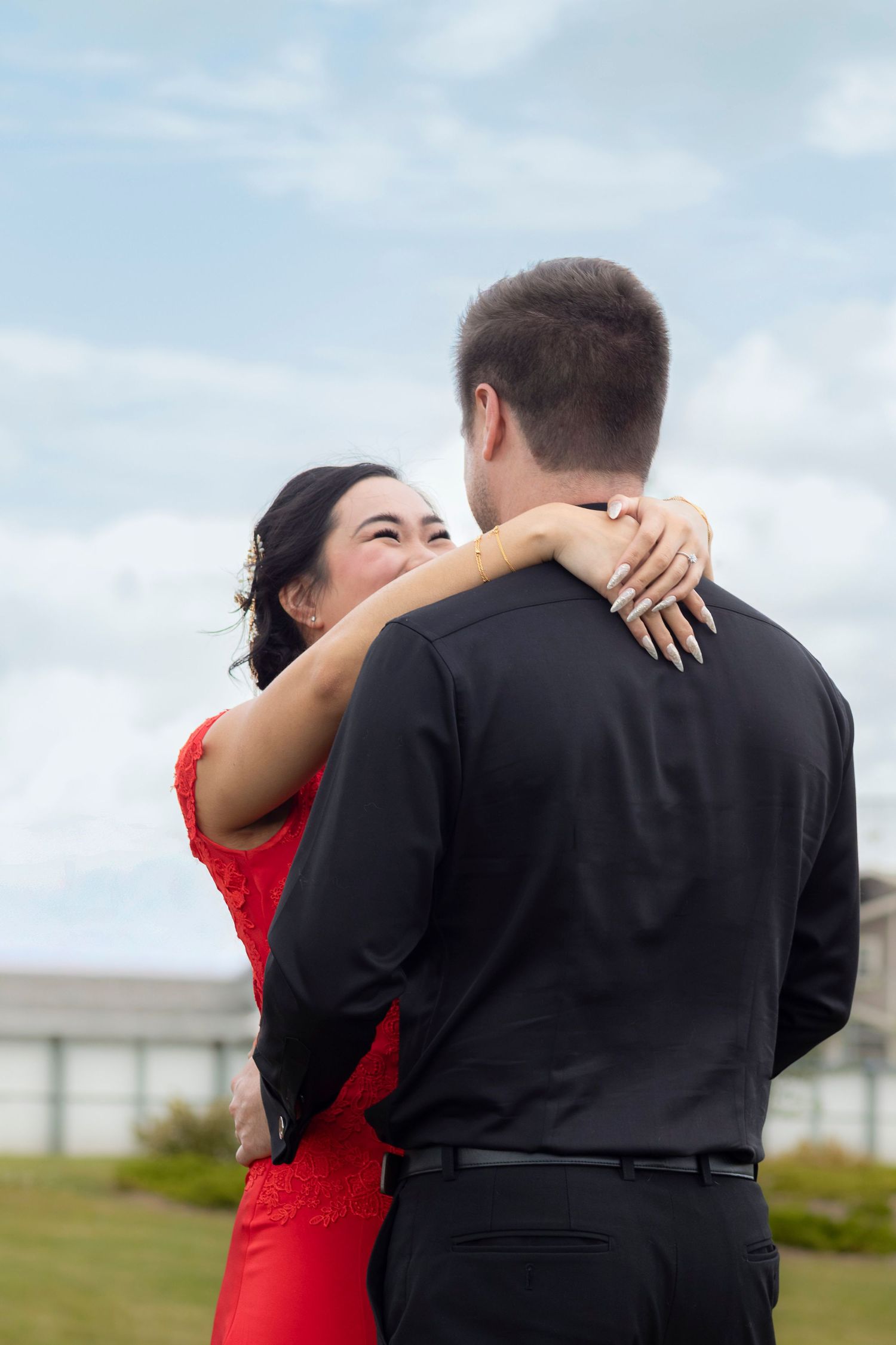 A couple in a red dress and black suit embrace romantically against a cloudy sky backdrop.