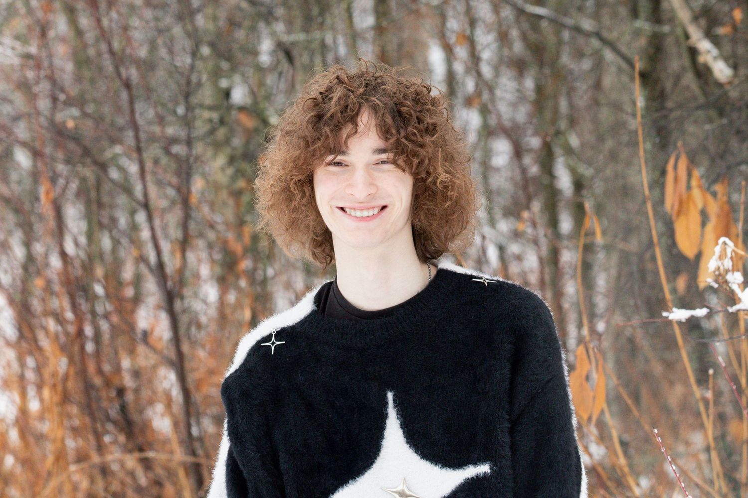 Smiling person with curly hair wearing a black and white sweater stands in front of snowy winter trees.