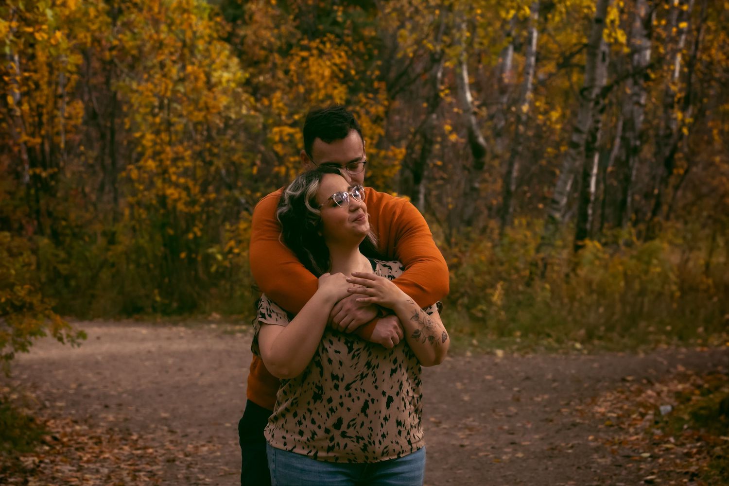 A couple embraces on a forest path surrounded by golden autumn foliage during sunset.