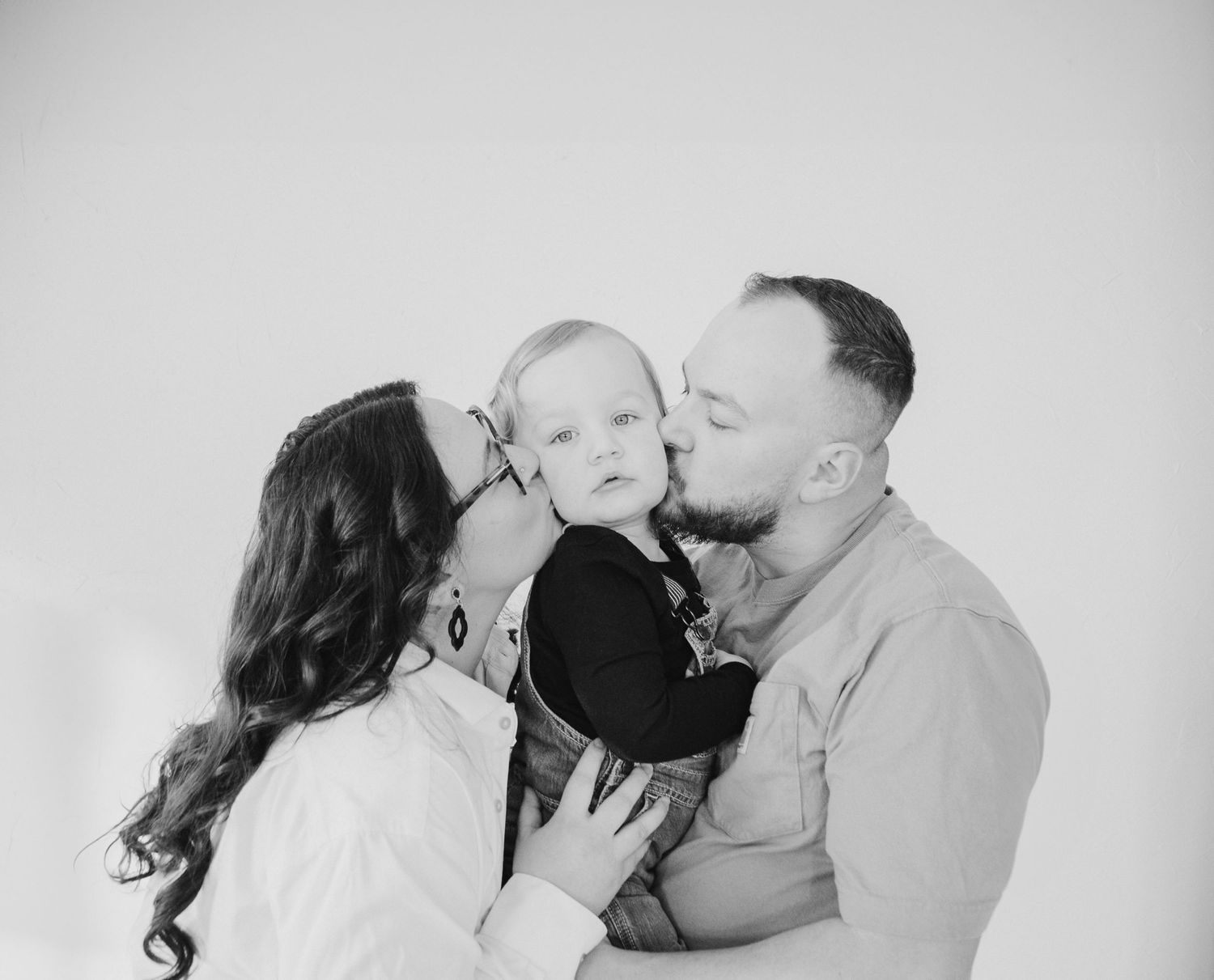 A heartwarming black and white family portrait shows parents lovingly kissing their small child from both sides.