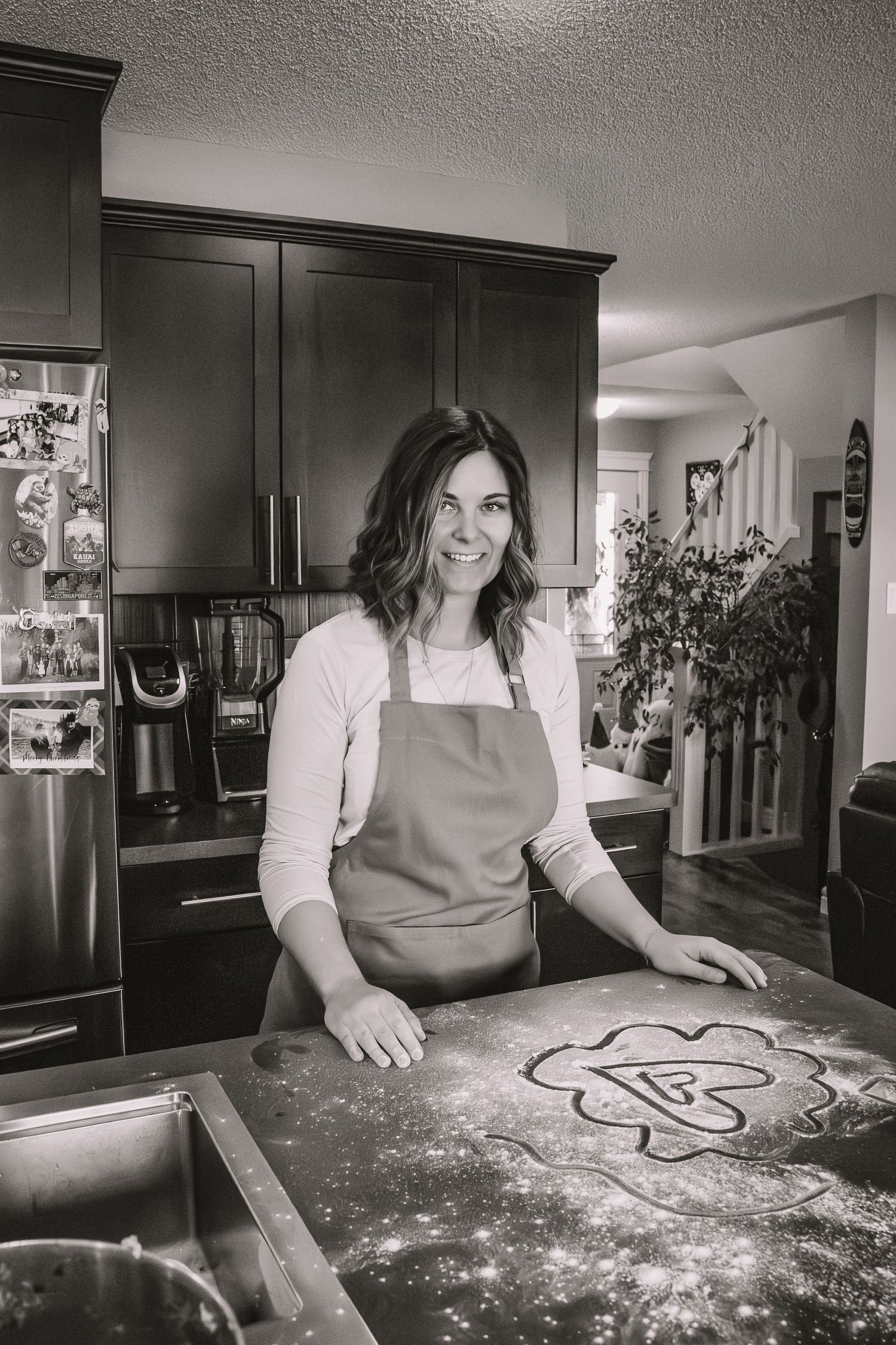 A baker in an apron draws a heart shape in flour on a dark countertop in a black and white kitchen scene.