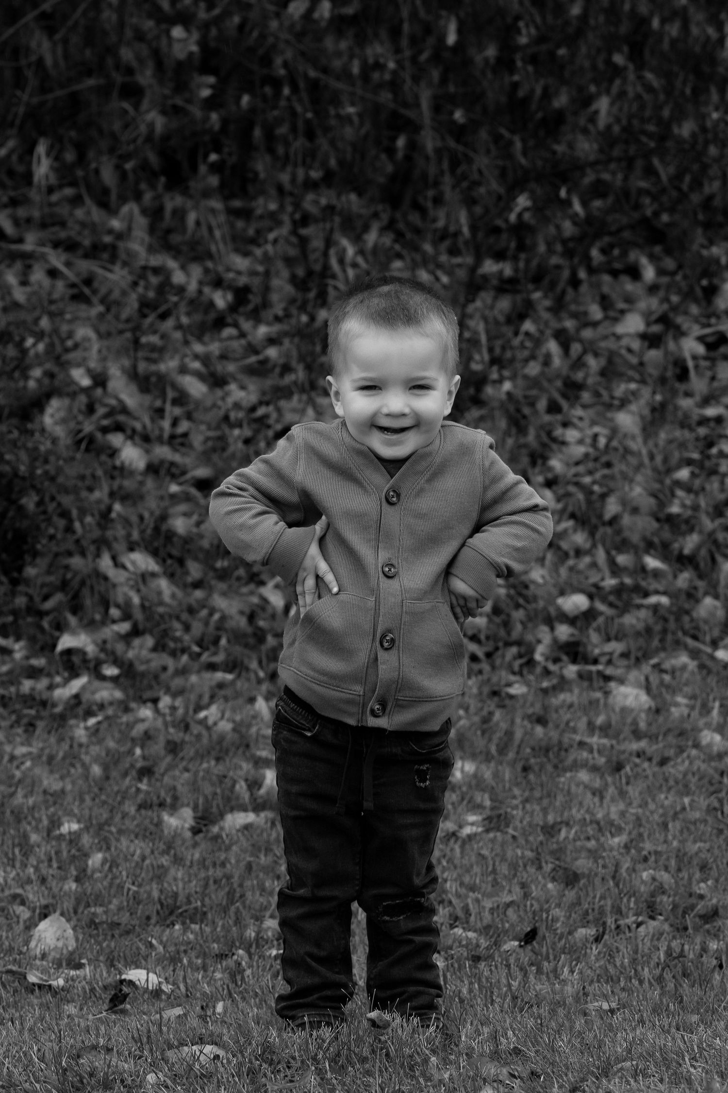 A young child stands on grass wearing a button-up cardigan and dark pants in this black and white photograph.