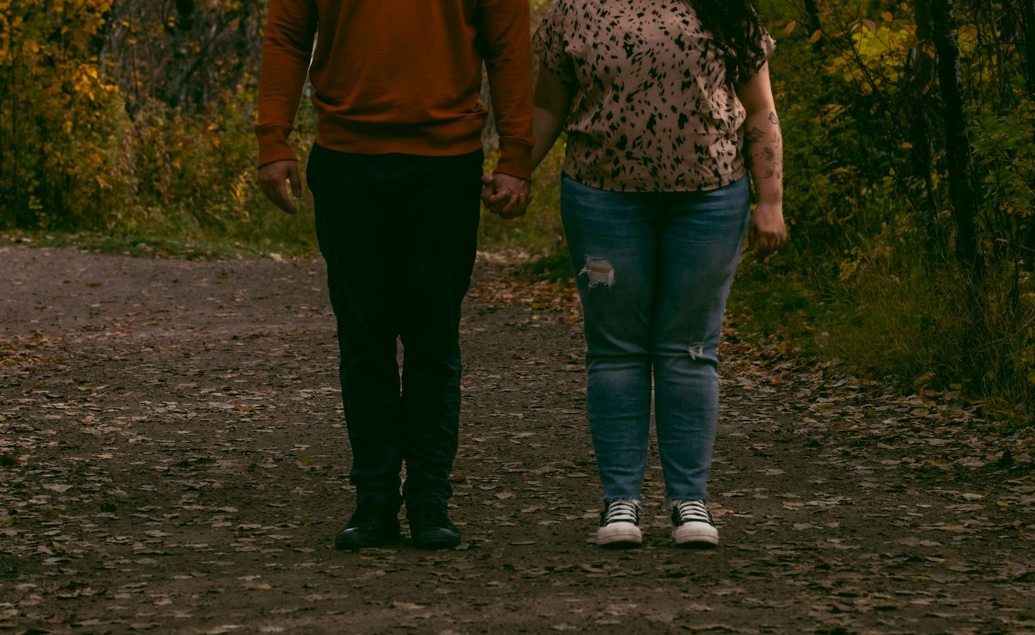 Two people standing together on a dirt path wearing casual clothing and jeans during autumn.