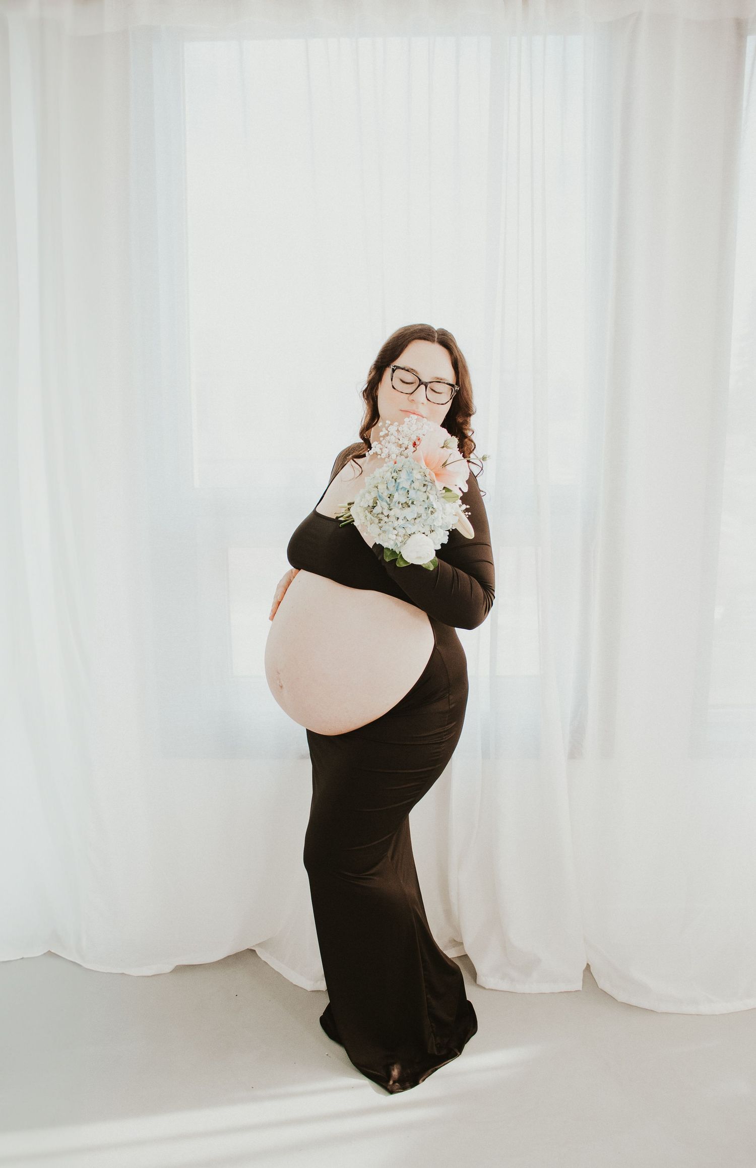 A pregnant woman in a long black dress holds white flowers during a maternity photoshoot against a light backdrop.