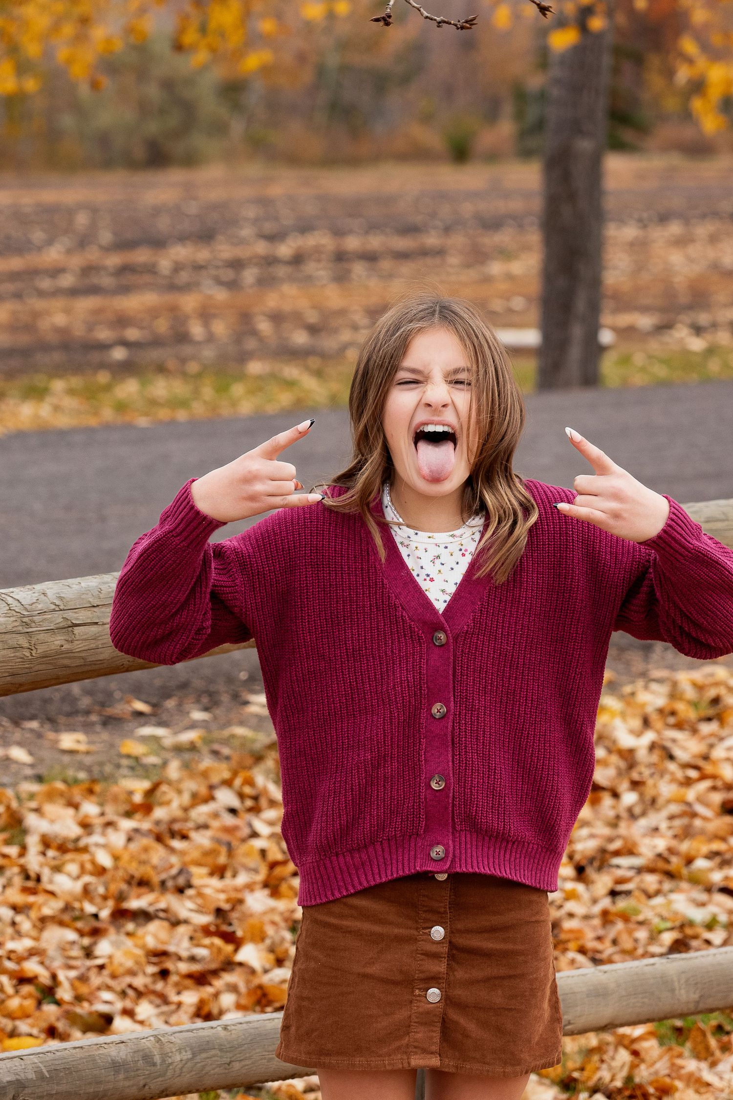 Person wearing burgundy cardigan and brown skirt makes rock and roll gesture in autumn scene with fallen leaves on ground.