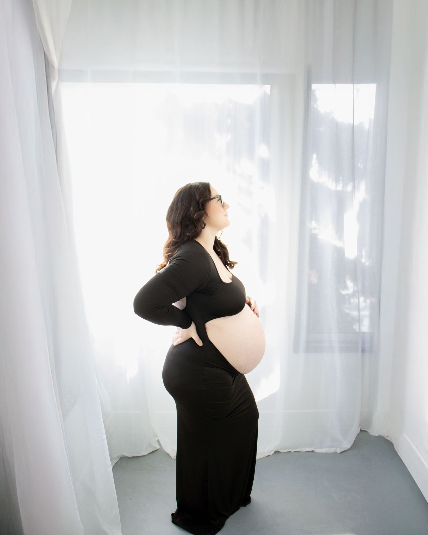 A pregnant woman in a long black dress stands in profile by sheer white curtains in soft natural lighting.
