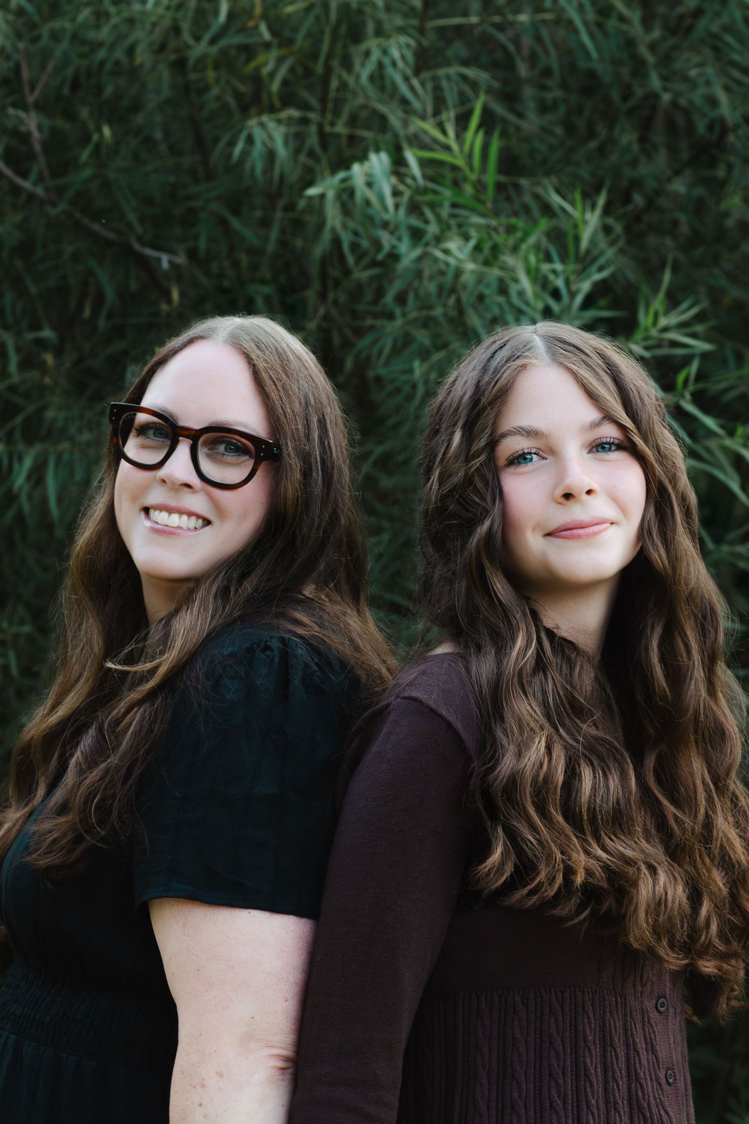 Two people in dark clothing share a playful moment while posing together against a green leafy background.