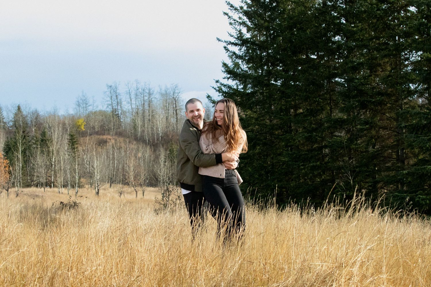 A couple shares a joyful embrace while standing in a golden field with evergreen trees and forest in the background.