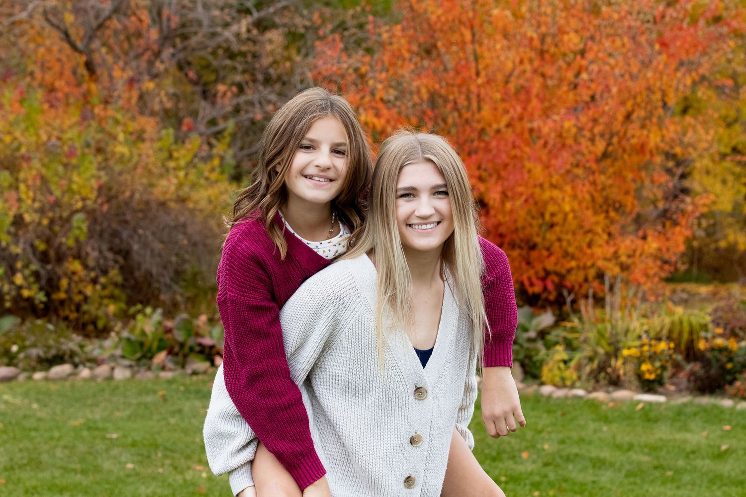 Two people wearing burgundy and white sweaters pose together against vibrant autumn foliage in a garden setting.