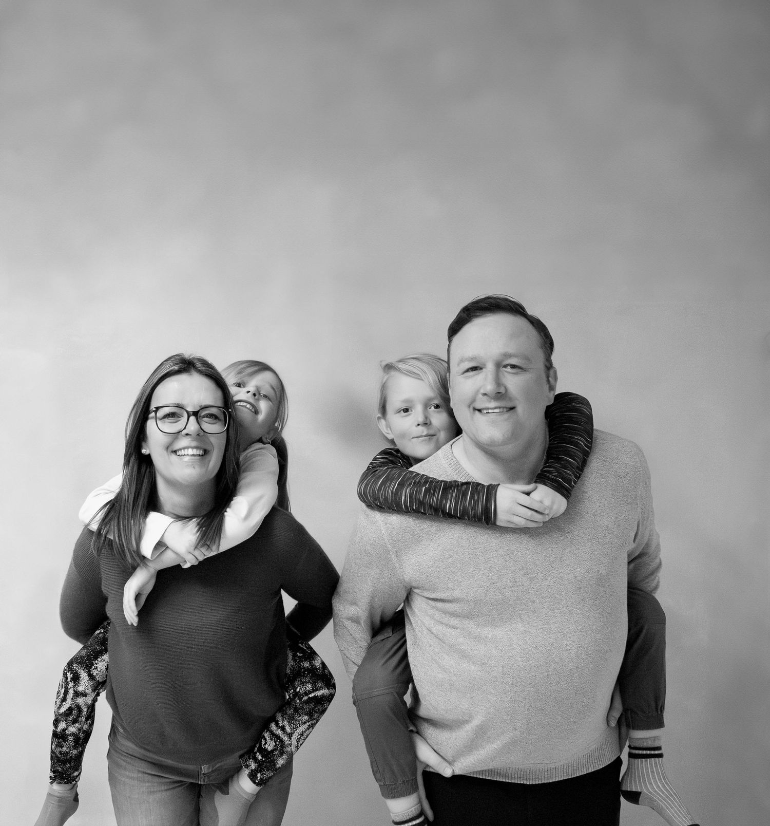 Black and white portrait of a family of four hugging and smiling together against a light background.