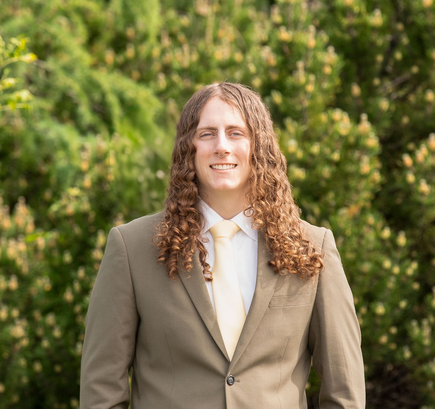 Portrait of person with curly hair wearing tan suit jacket and white shirt outdoors.