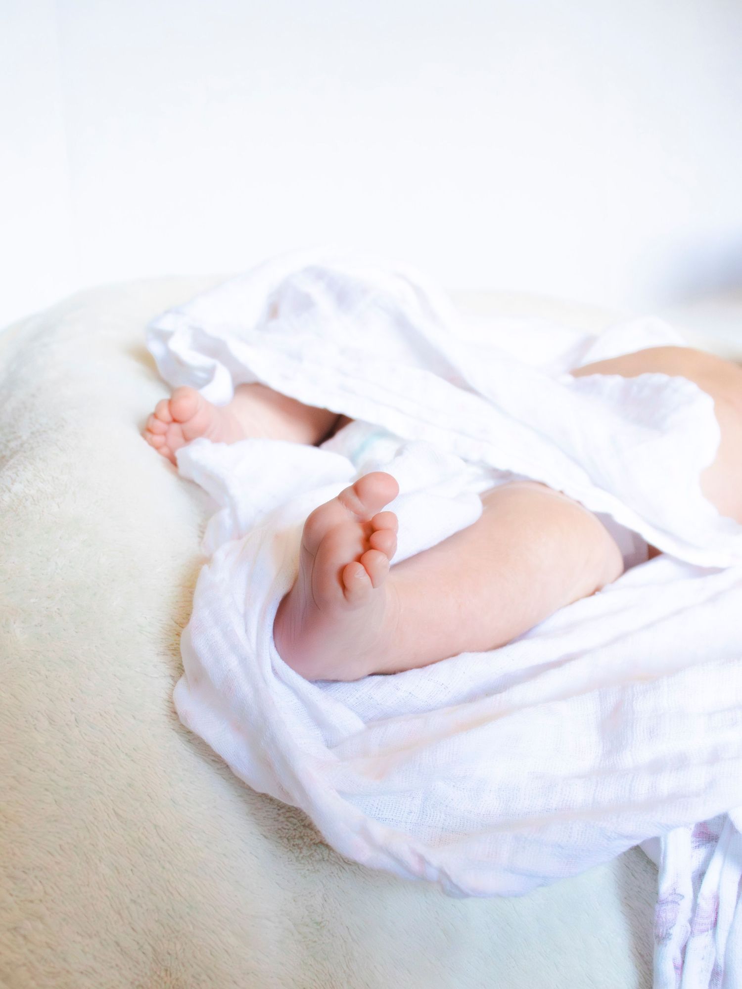 A small baby's feet wrapped in soft white fabric are delicately positioned against a light cream-colored background.