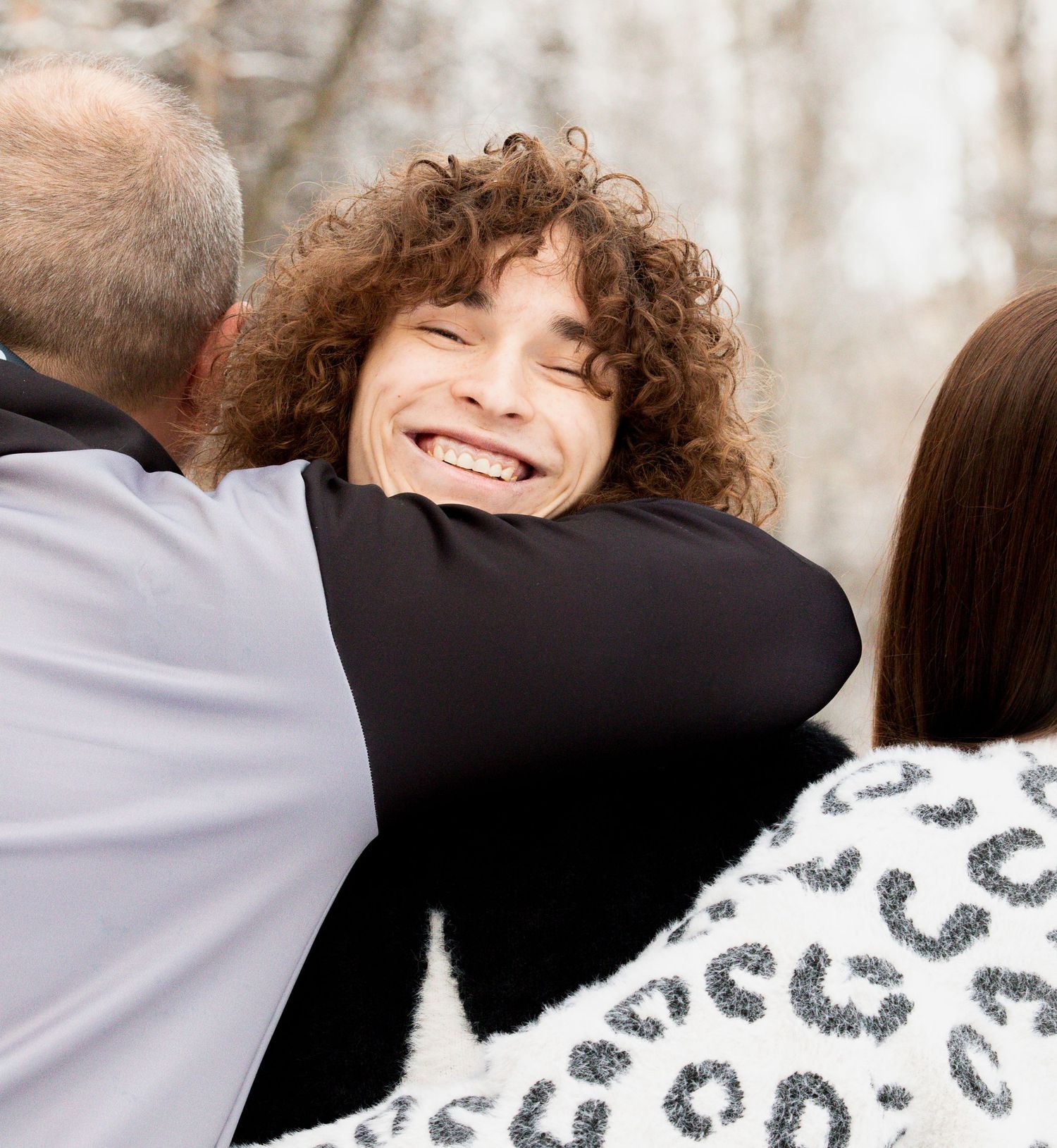 Person with curly hair smiling warmly while being embraced in a group hug outdoors in winter.