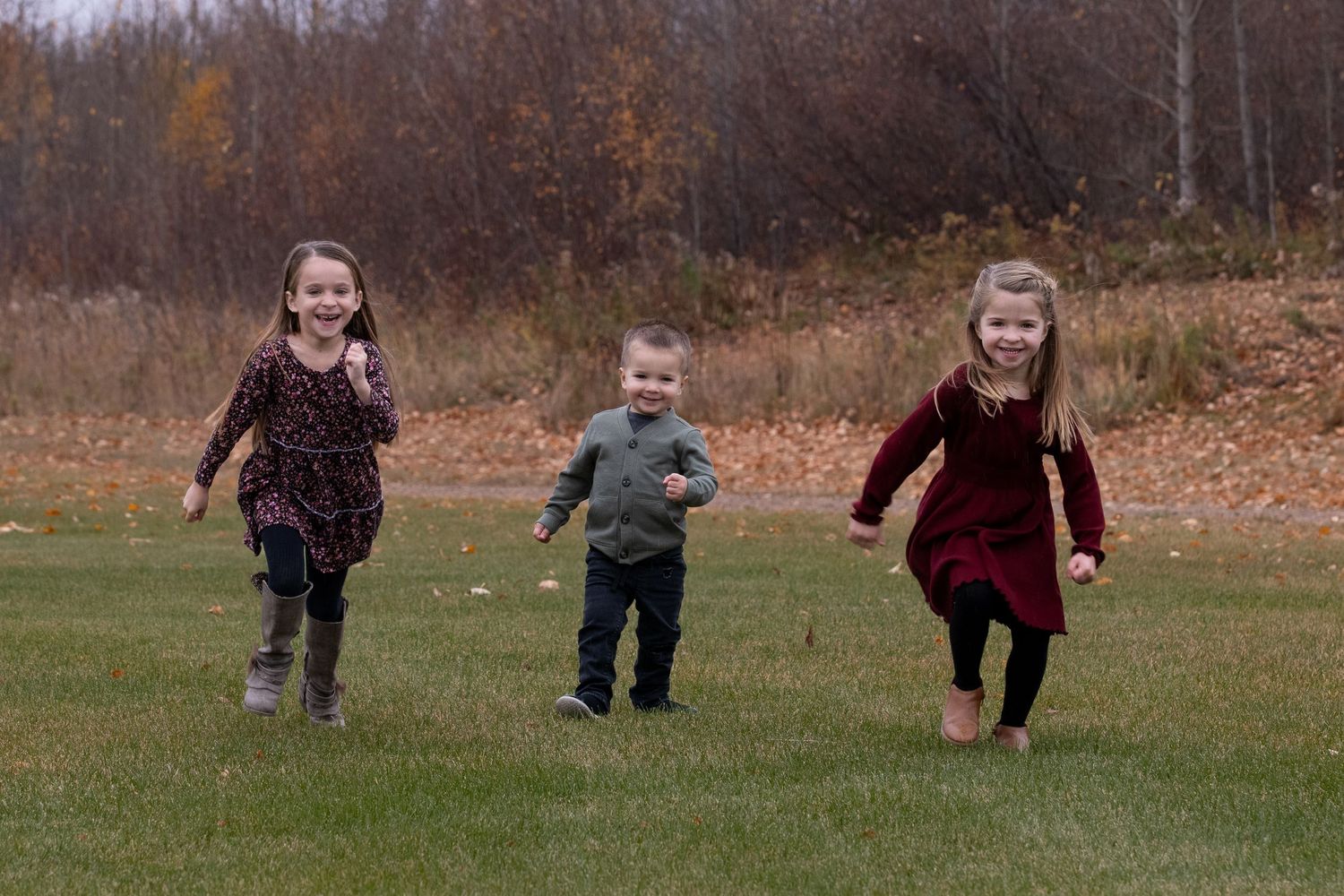 Three children run and play together on a grassy field during autumn with trees in the background.