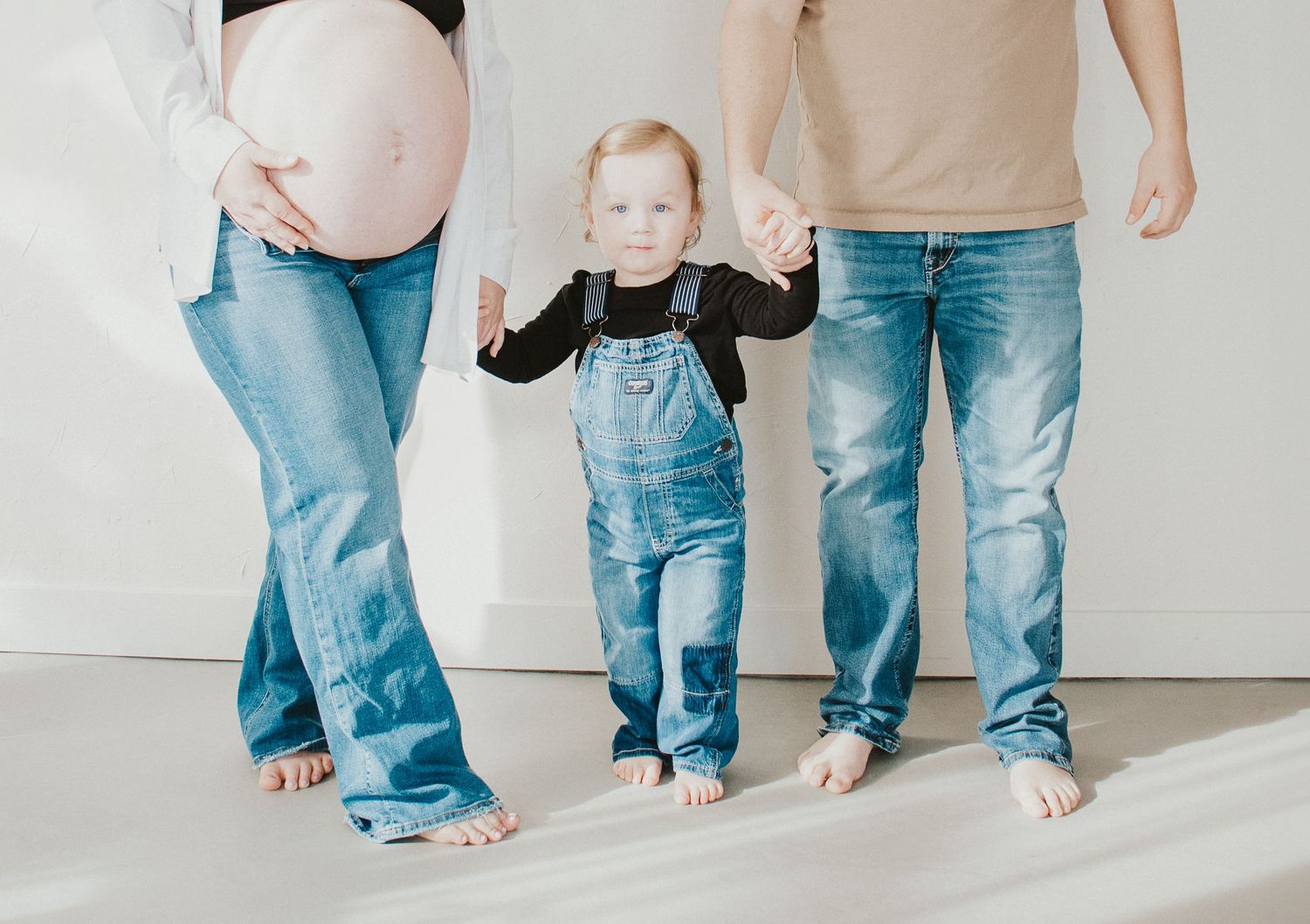 A small child wearing overalls stands between two adults in jeans against a white backdrop.