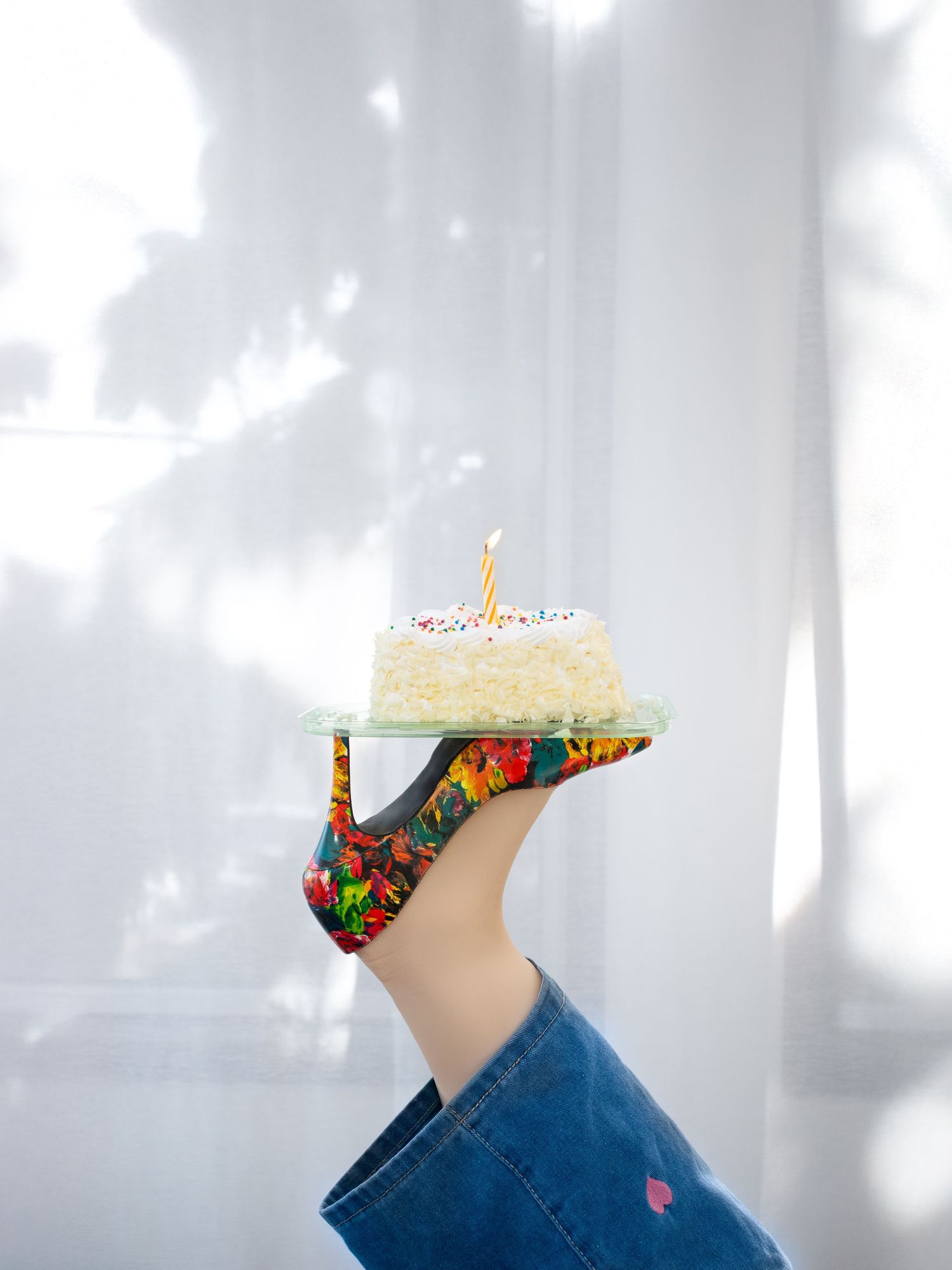 A photography website photo of a foot wearing a  floral high heel shoe that holds up a small white birthday cake with a single candle.