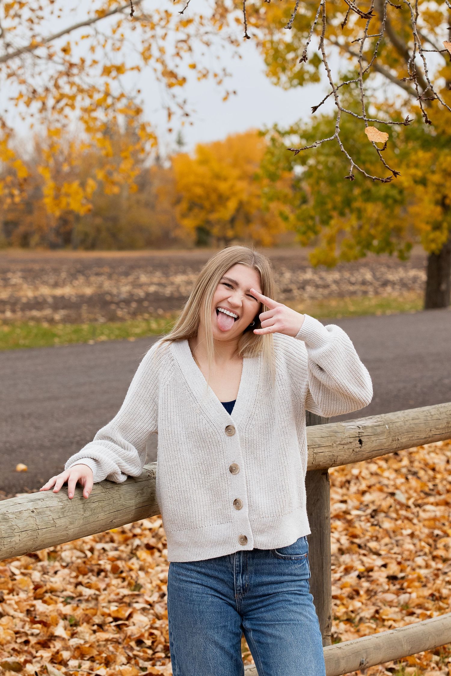 Cheerful person in cream sweater and jeans leaning against wooden fence surrounded by autumn leaves and golden trees.