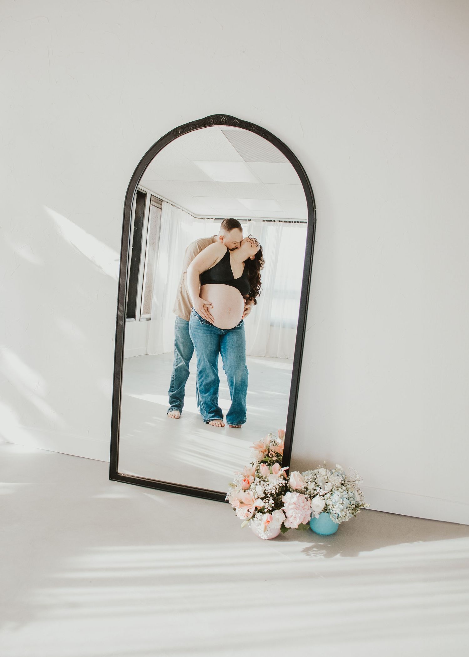 Mirror reflection shows a maternity moment with flowers and a decorative vase placed at the bottom of an arched frame.