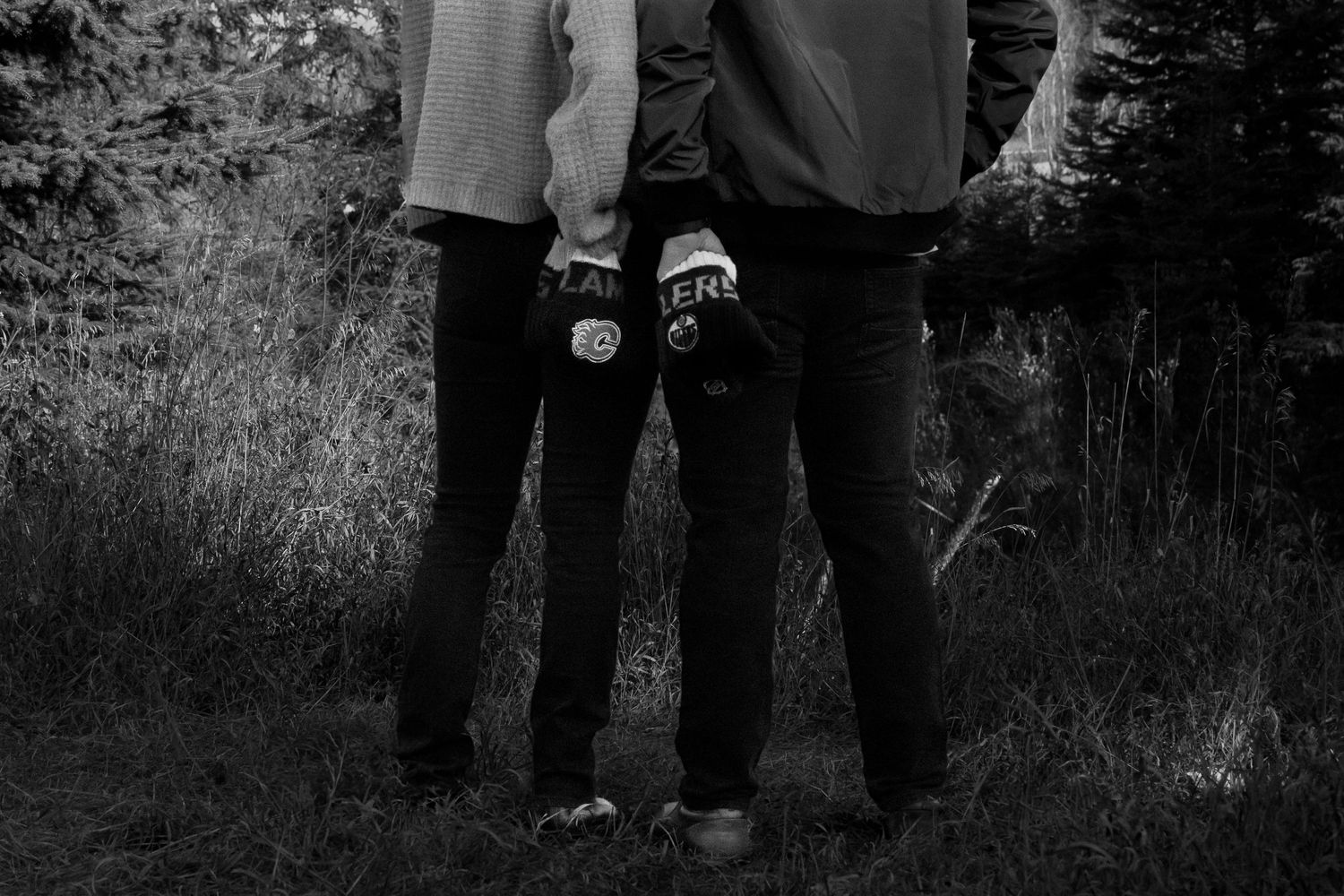 Two people stand closely together in a black and white photo showing hockey toques of opposing teams hanging out their pants back pockets